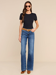 A woman with long hair stands against a plain white background, wearing sunglasses, a black short-sleeve shirt, brown heeled shoes, and the Clayton Wide-Leg Jean in a high-rise blue wash with a brown belt; one hand rests in her pocket.