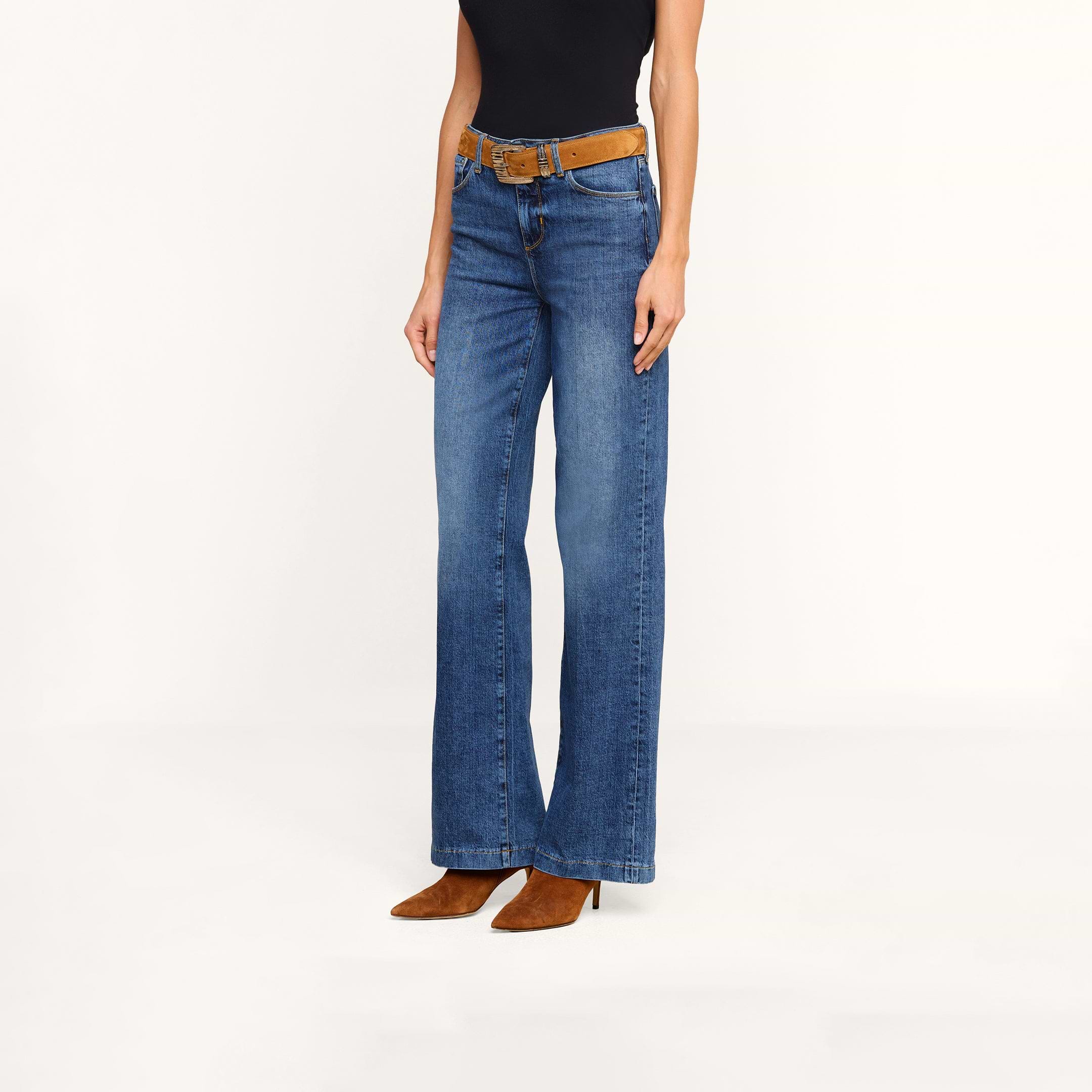 Person wearing the Clayton Wide-Leg Jean in blue stretch denim, styled with a brown belt, pointed brown boots, and a sleeveless black top, standing against a plain white background.