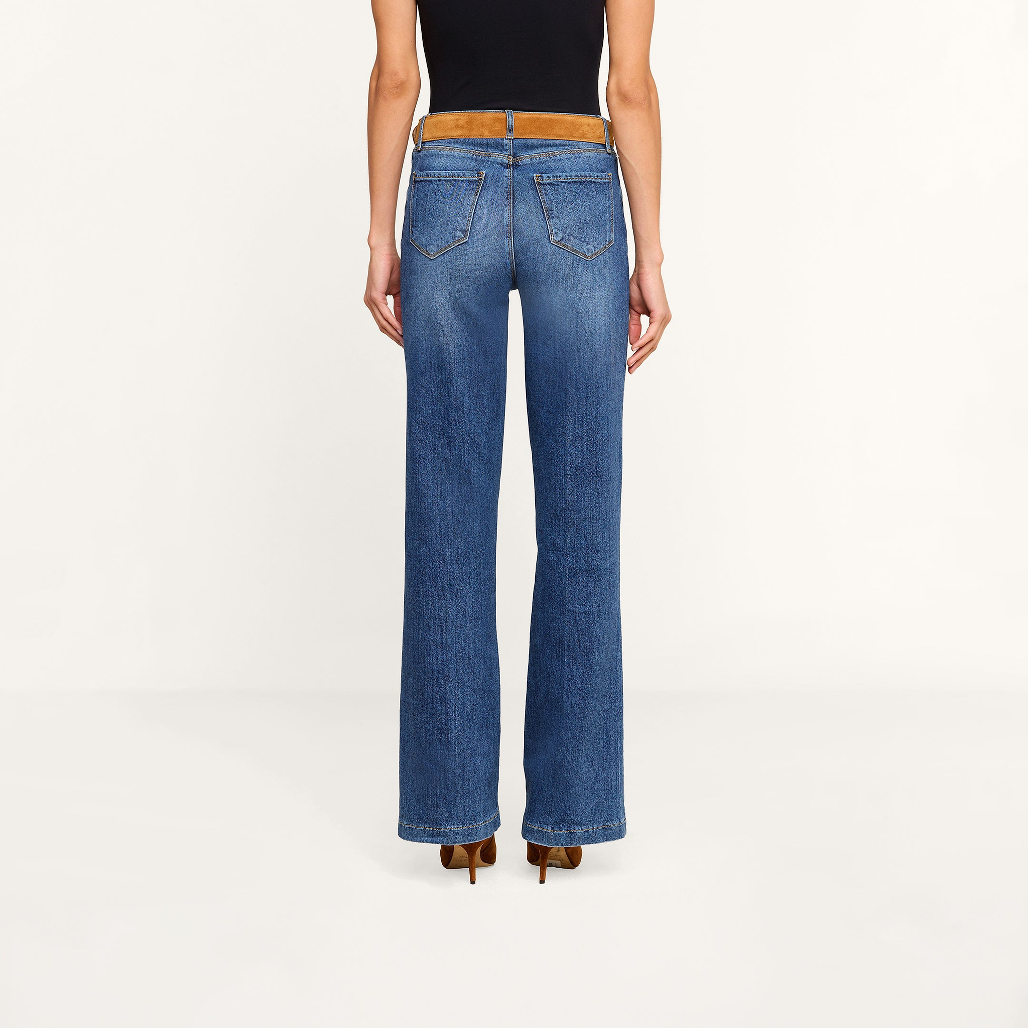 A person wearing the Clayton Wide-Leg Jean in blue, paired with a black sleeveless top, brown belt, and brown heeled shoes, is standing with their back to the camera against a plain white background.