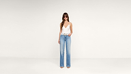 Woman stands against a plain white background wearing sunglasses, a white sleeveless top, light blue high-waisted wide-leg jeans, and open-toe heels. She poses with one hand in her pocket.