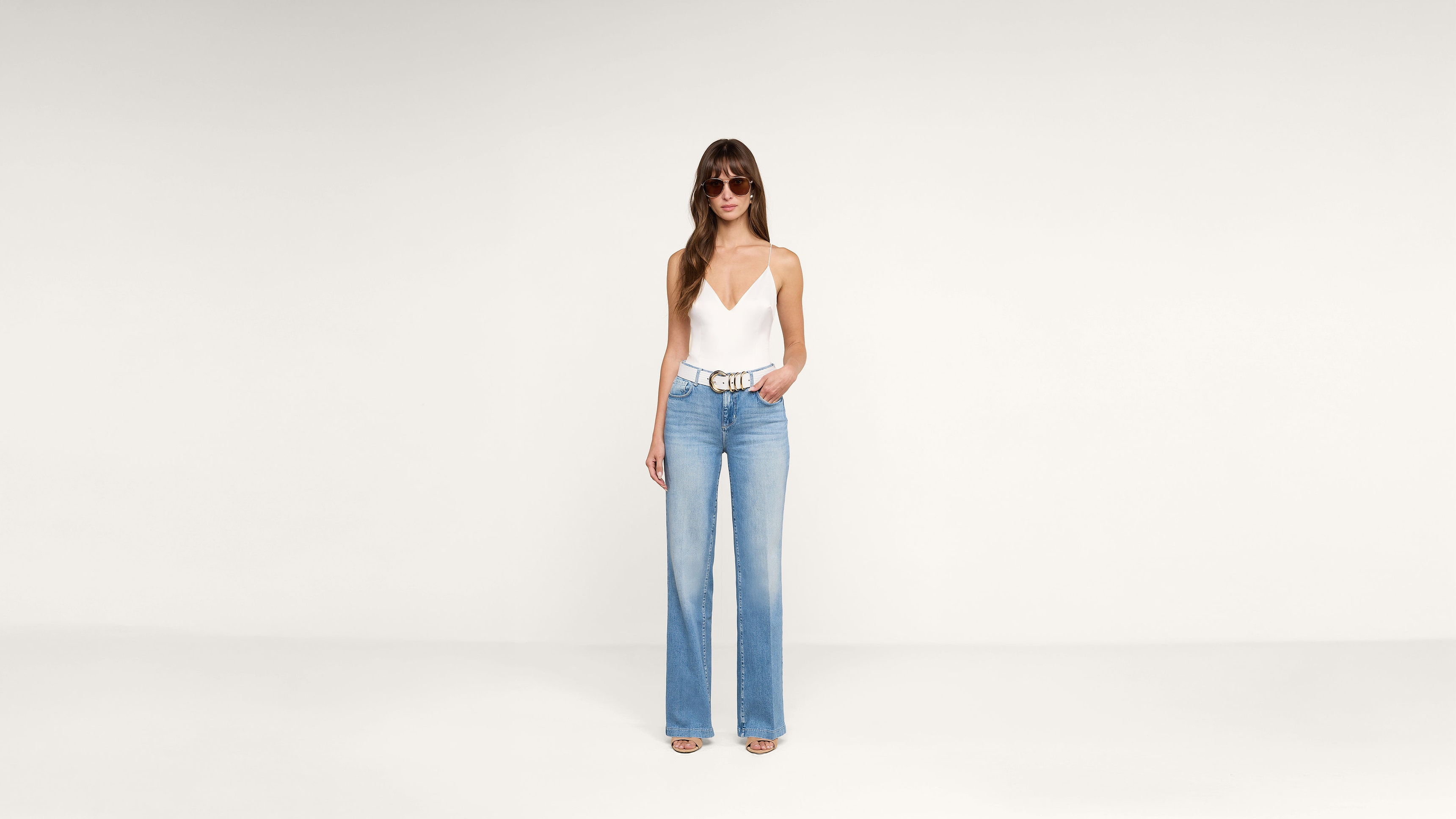 Woman stands against a plain white background wearing sunglasses, a white sleeveless top, light blue high-waisted wide-leg jeans, and open-toe heels. She poses with one hand in her pocket.