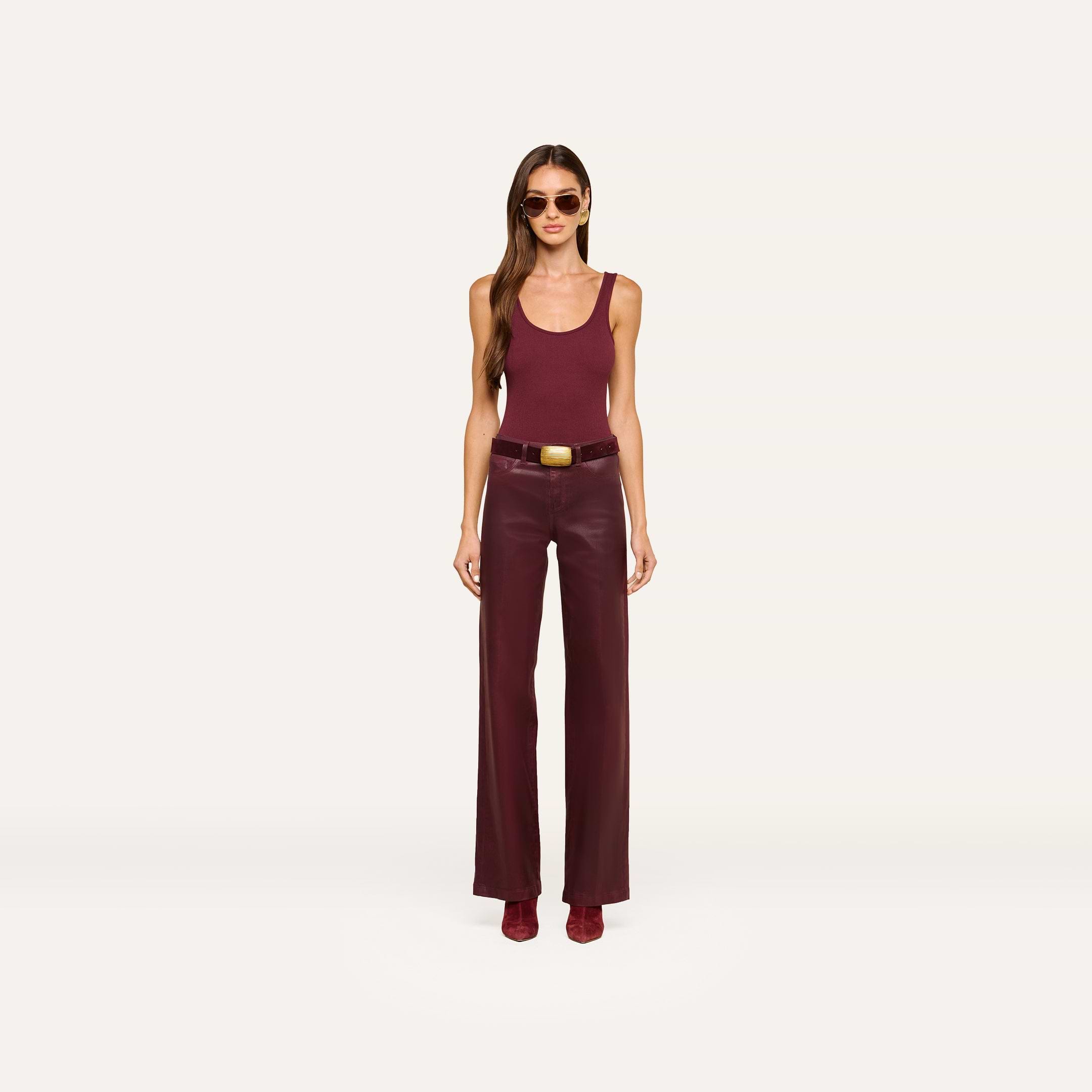A woman wearing sunglasses, a sleeveless burgundy top, the Clayton Coated Wide-Leg Jean with a contoured waistband and belt, and matching shoes stands against a plain white background.