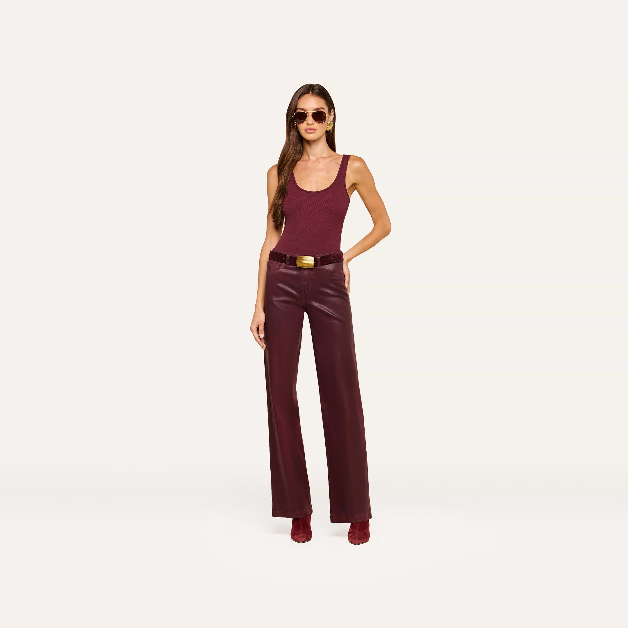 A woman wearing sunglasses, a sleeveless maroon top, the Clayton Coated Wide-Leg Jean with a contoured waistband, a belt with a gold buckle, and matching boots stands against an off-white background.