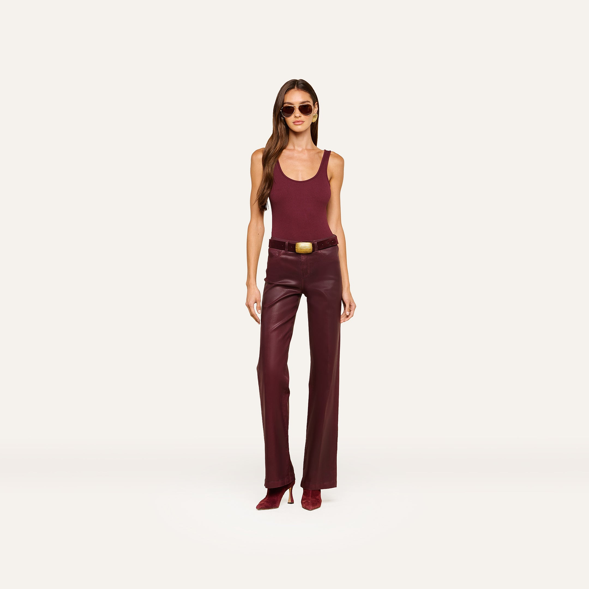 A woman wearing sunglasses, a sleeveless maroon top, the Clayton Coated Wide-Leg Jean with a contoured waistband and belt in dark maroon, and maroon heeled shoes, stands against a plain light background.