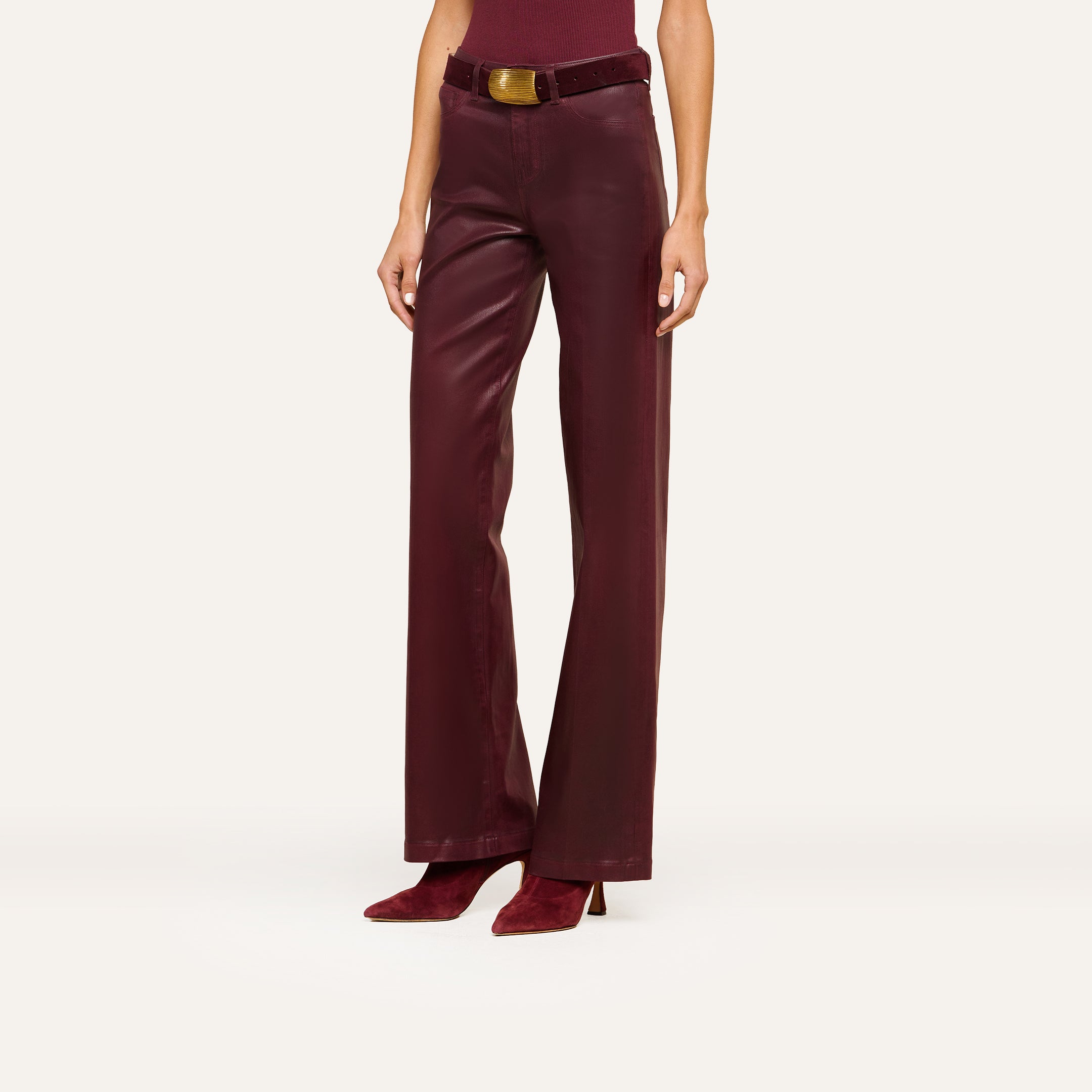 A person models the Clayton Coated Wide-Leg Jean, featuring a high waist, wide-leg silhouette, contoured waistband, and matching belt in burgundy leather. They pair the jeans with pointed burgundy heeled shoes. The top of their head is not visible.