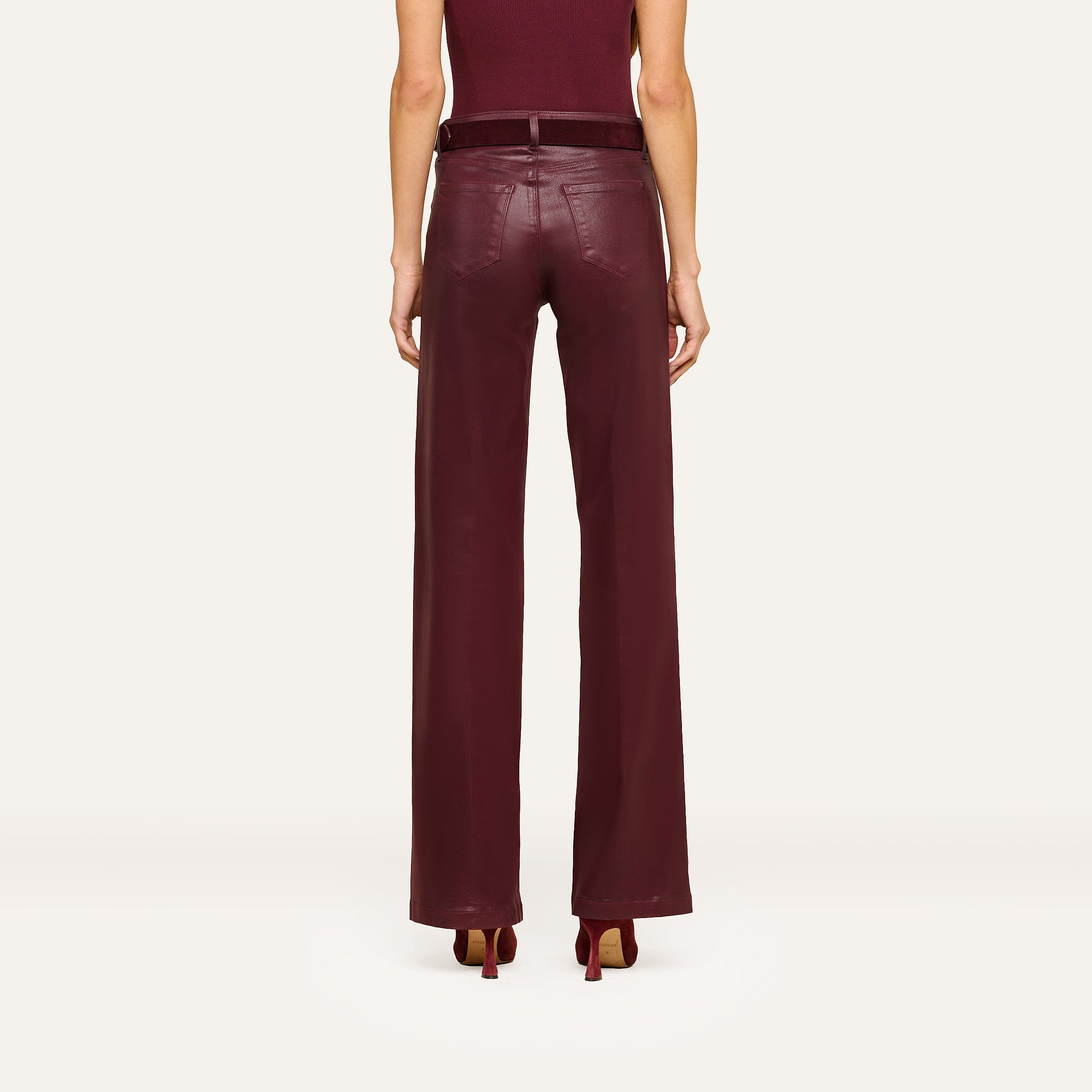 A person stands facing away, wearing the Clayton Coated Wide-Leg Jean featuring a contoured waistband in a rich burgundy hue, paired with a matching belt and heels. The background is plain and light-colored.