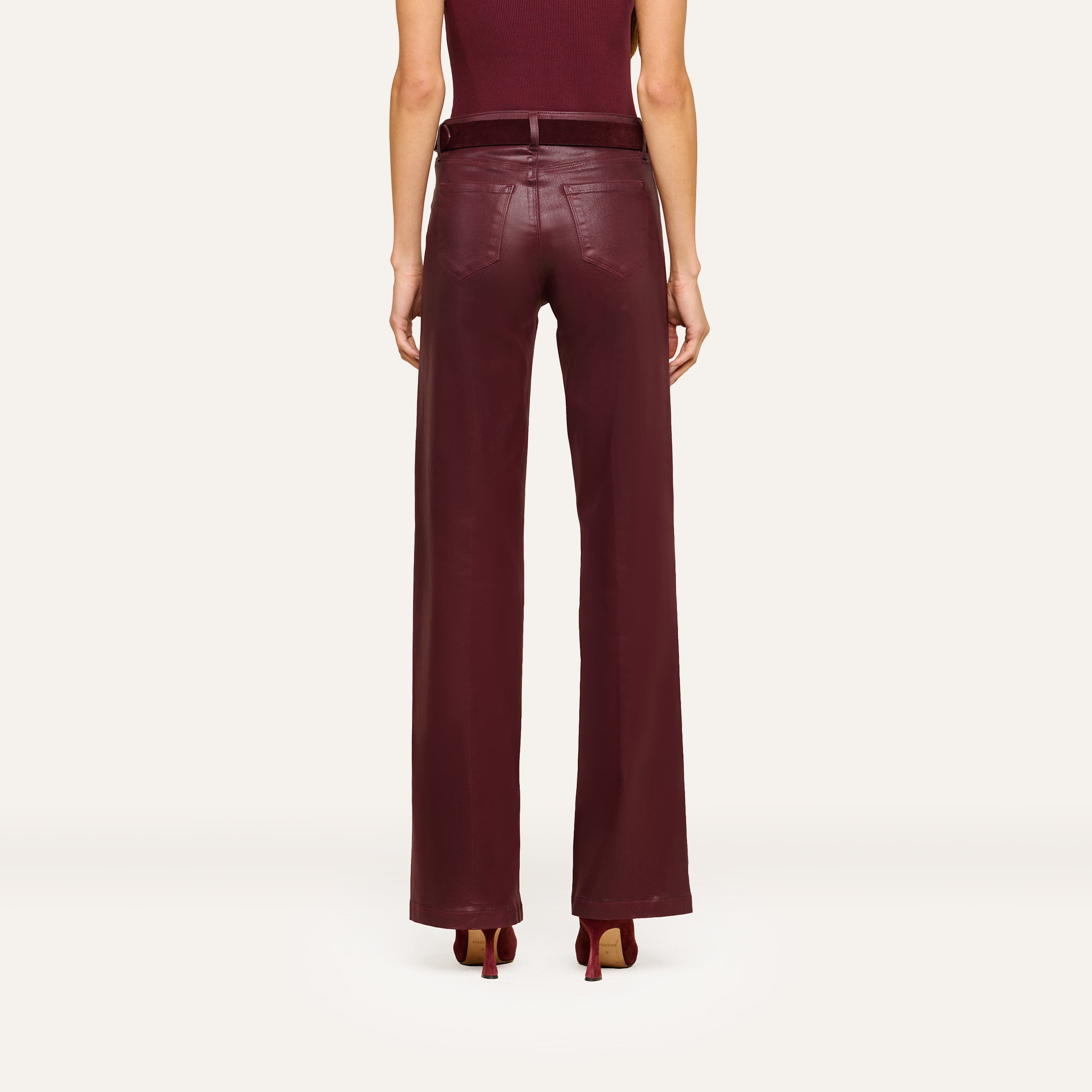 A person stands facing away, wearing the Clayton Coated Wide-Leg Jean featuring a contoured waistband in a rich burgundy hue, paired with a matching belt and heels. The background is plain and light-colored.