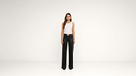 A woman stands against a plain white background, wearing a white sleeveless top, black wide-leg pants with a gold belt buckle, and black pointed shoes.