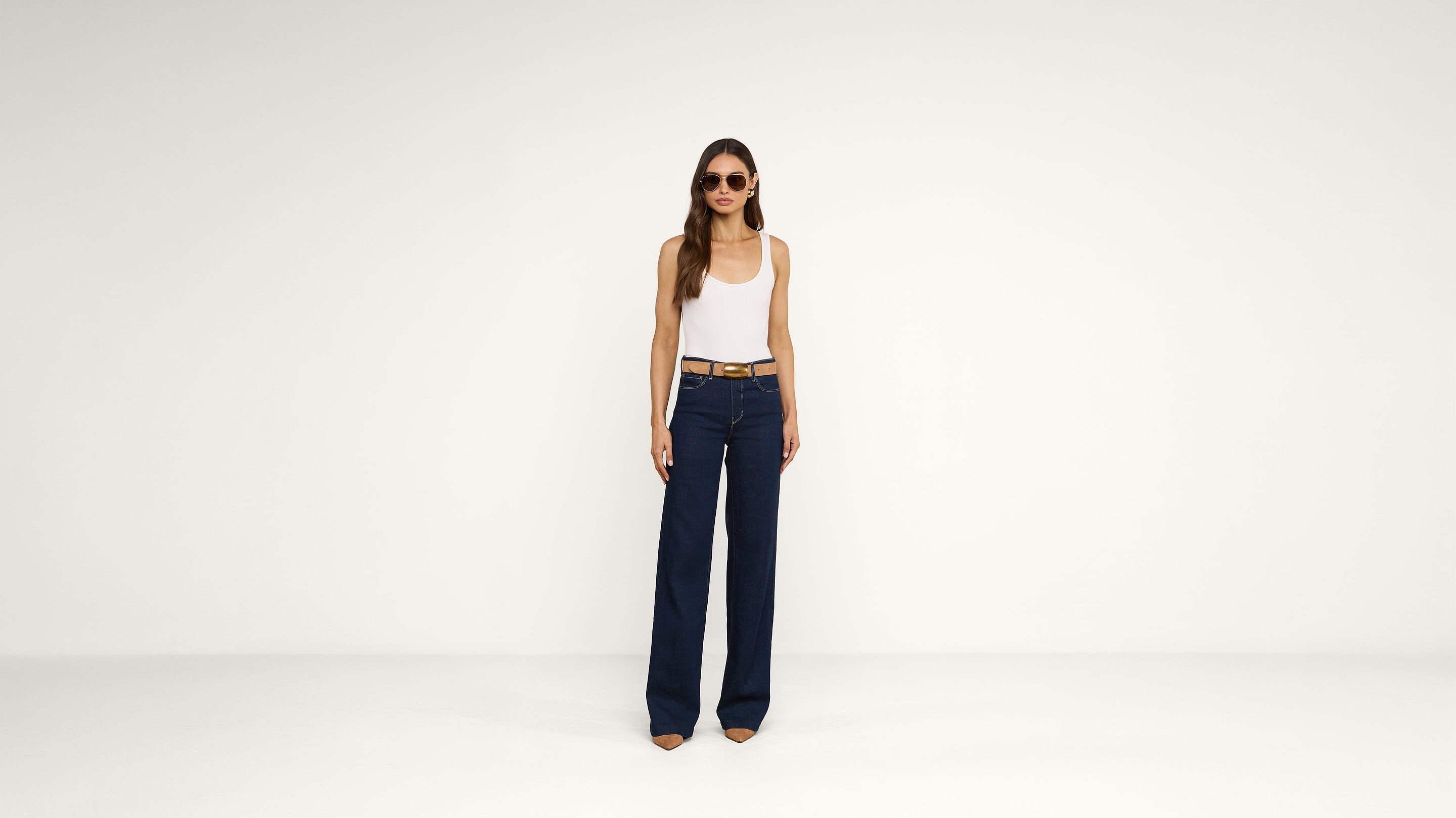 A woman wearing sunglasses, a white tank top, a brown belt, and wide-leg dark blue jeans stands against a plain white background.