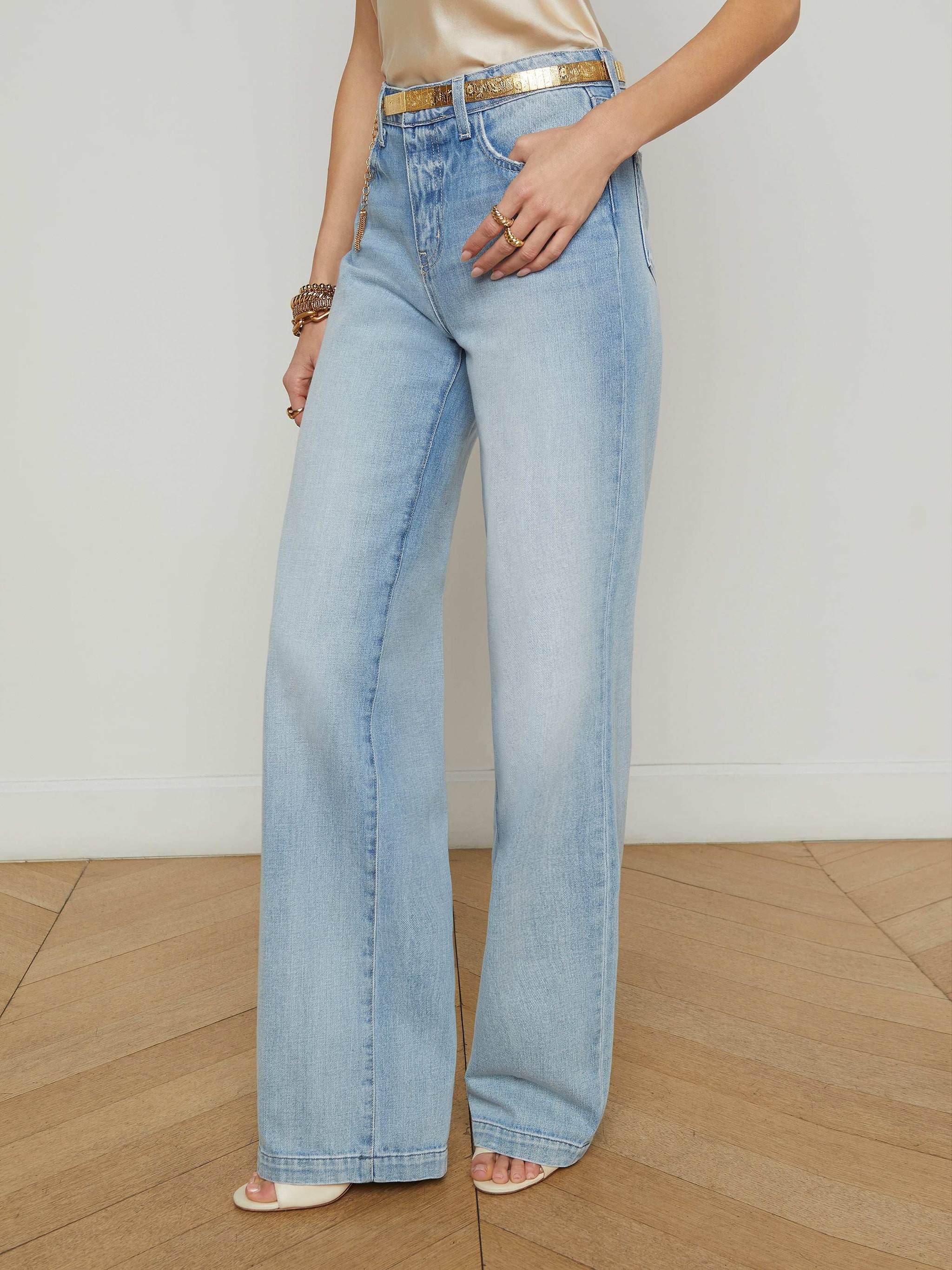 A woman wears the Clayton Wide-Leg Jean in a light wash with a gold belt and beige heels, standing with one hand in her pocket against a plain background.
