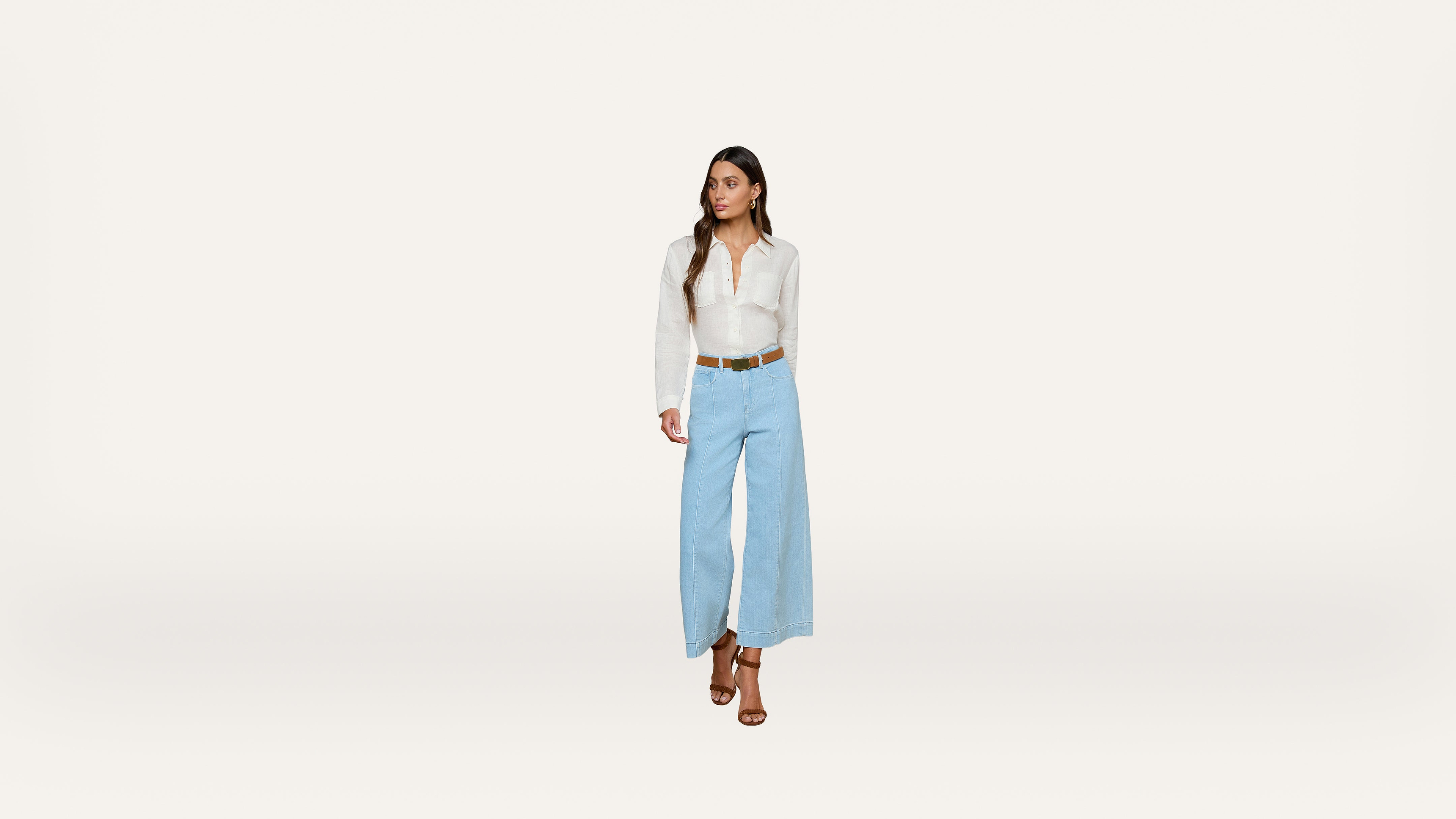 A woman wearing a white long-sleeve shirt, light blue wide-leg pants, a brown belt, and brown heeled sandals stands against a plain white background, looking to her left.