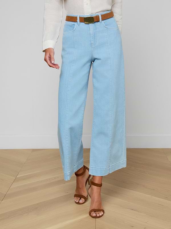 A person wearing the Houston Cropped Wide-Leg Jean in light blue stretch denim, styled with a brown belt, a white shirt, and brown sandals, stands against a plain background.