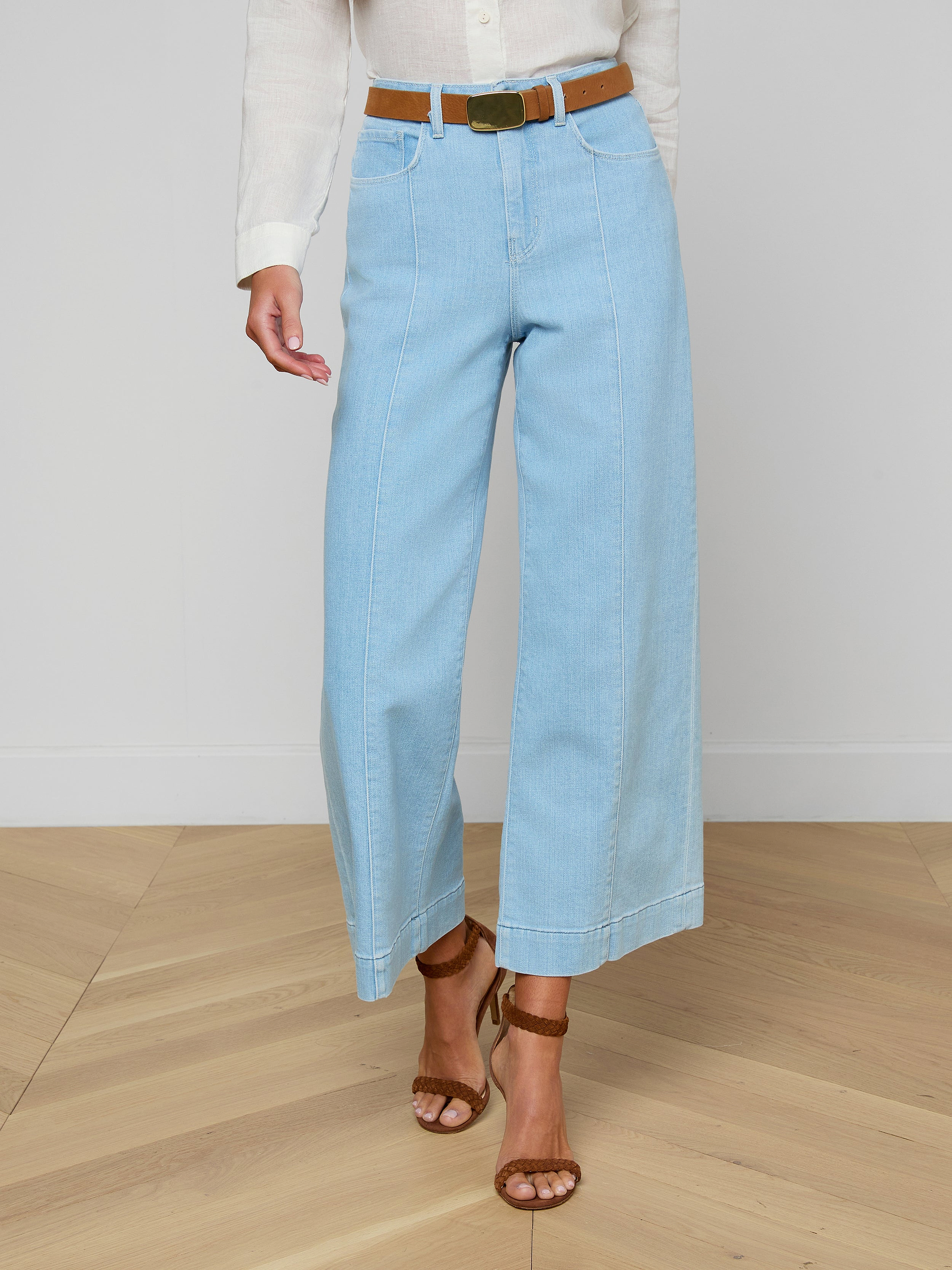 A person wearing the Houston Cropped Wide-Leg Jean in light blue stretch denim, styled with a brown belt, a white shirt, and brown sandals, stands against a plain background.