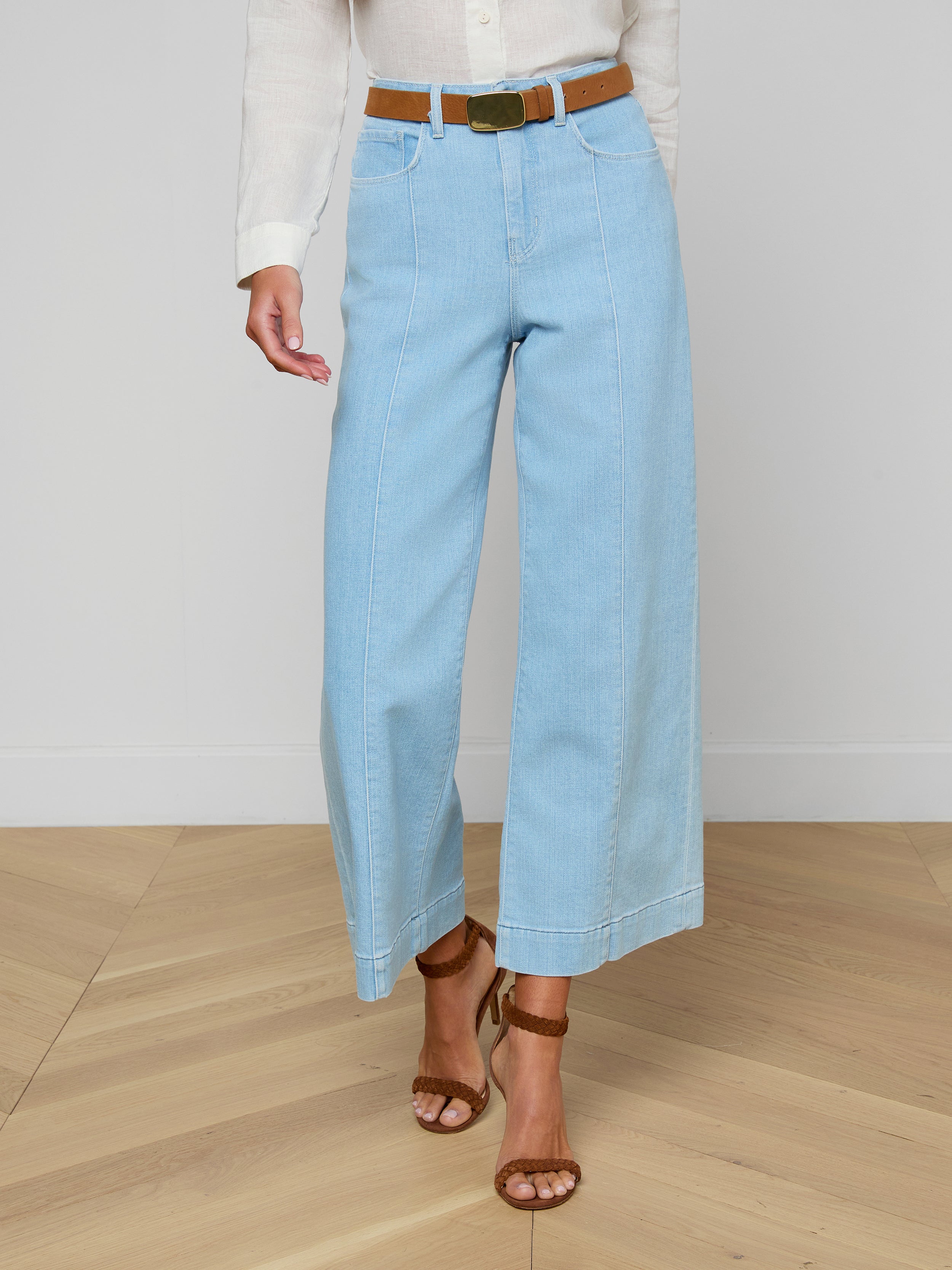 A person wearing the Houston Cropped Wide-Leg Jean in light blue stretch denim, styled with a brown belt, a white shirt, and brown sandals, stands against a plain background.