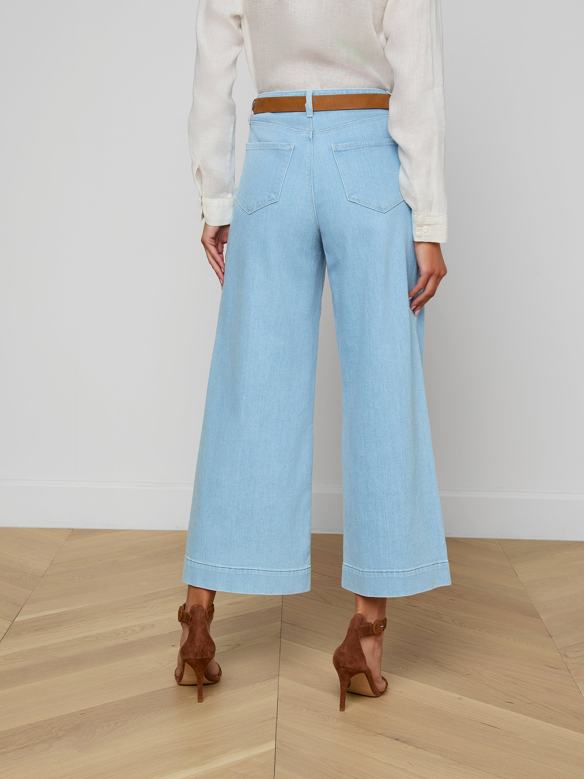 A woman is shown from the back against a plain background, wearing the Houston Cropped Wide-Leg Jean in light blue stretch denim, styled with a white long-sleeve top, a brown belt, and brown high-heeled shoes.