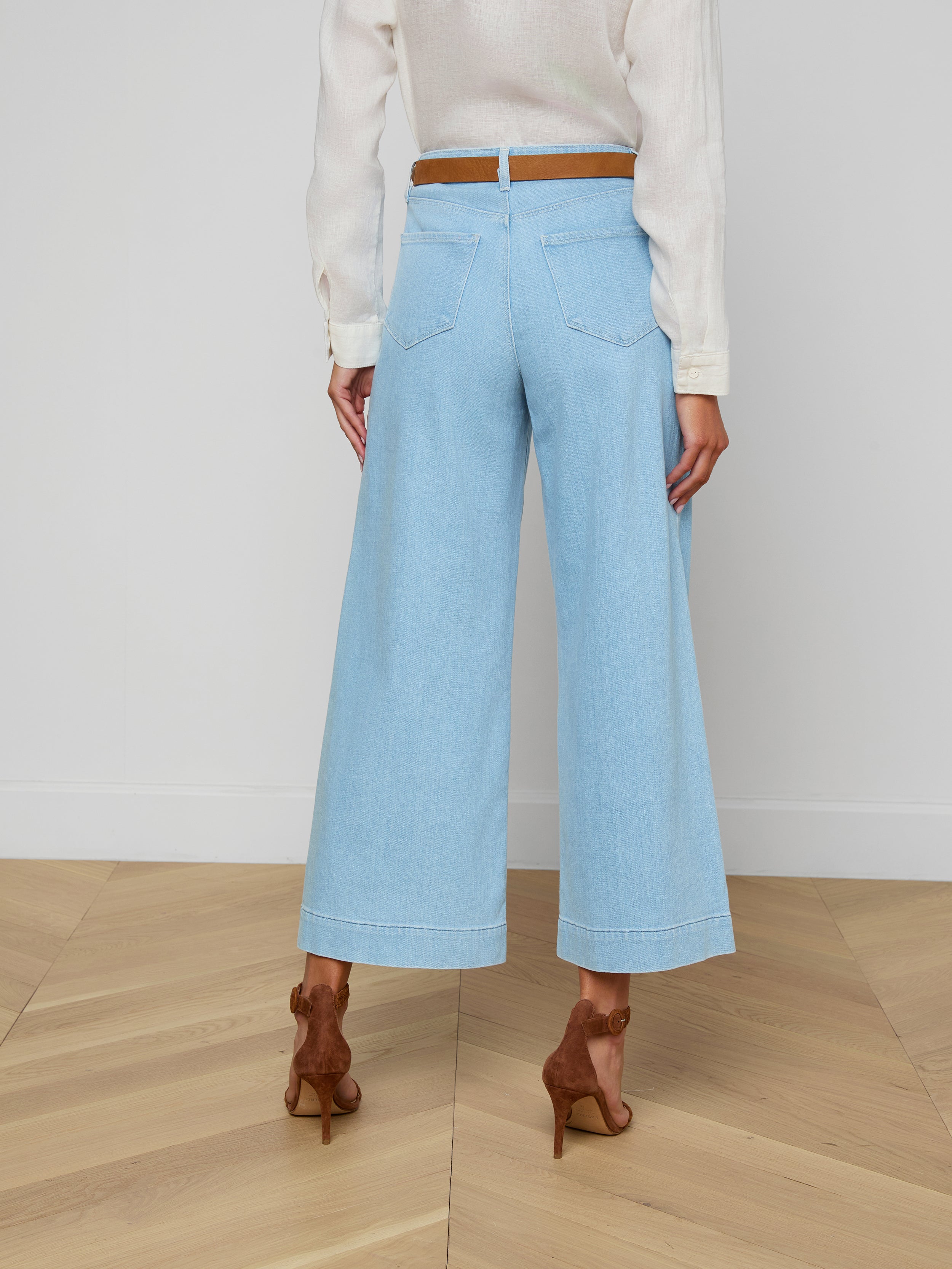 A woman is shown from the back against a plain background, wearing the Houston Cropped Wide-Leg Jean in light blue stretch denim, styled with a white long-sleeve top, a brown belt, and brown high-heeled shoes.