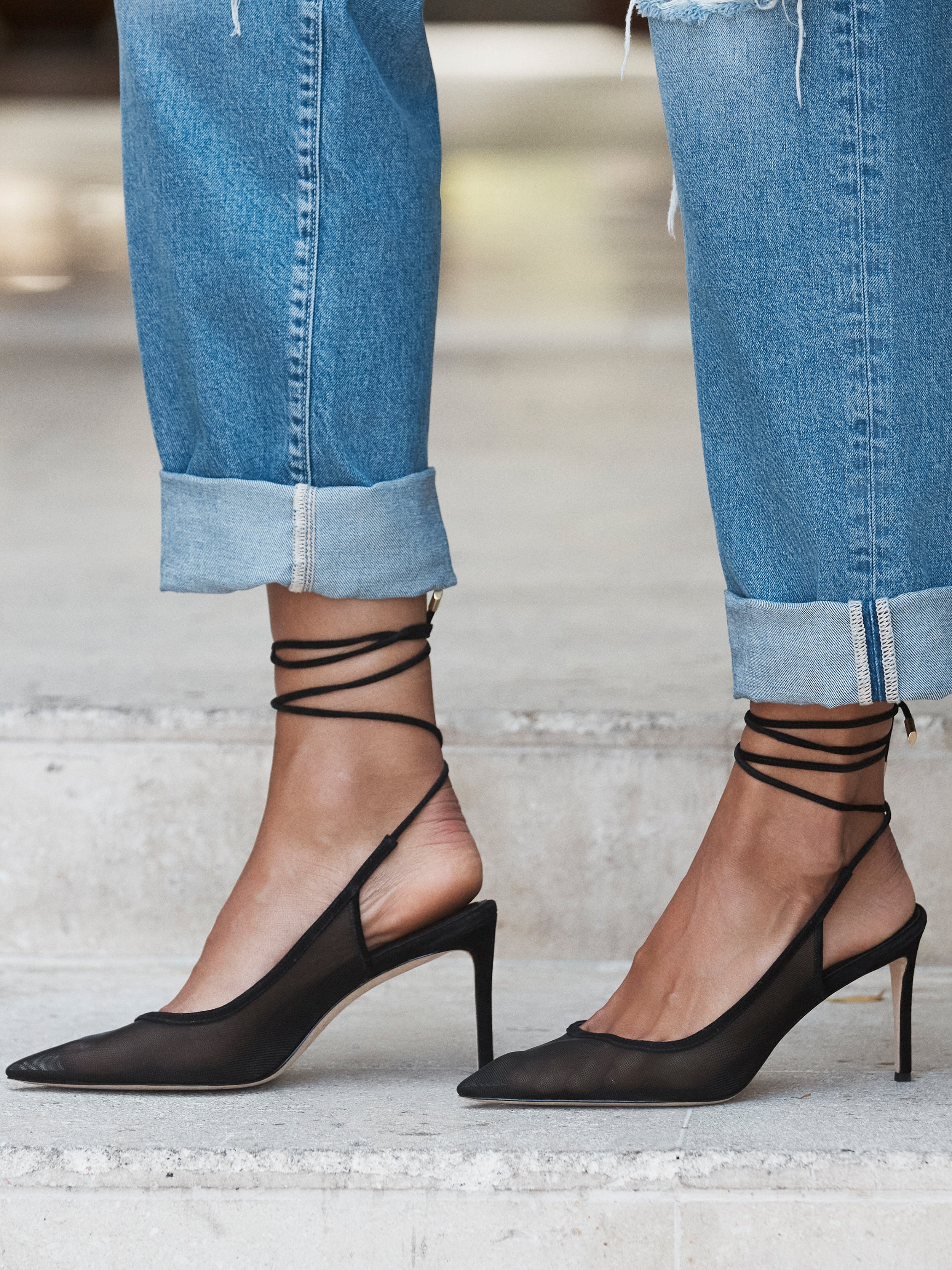 A person in blue cuffed jeans and black pointed-toe heels with ankle wrap straps stands on light-colored stone steps.