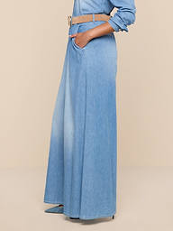 A person is wearing the Lorenza Palazzo Jean in light blue featherweight chambray denim, styled with a wide tan belt, hands in pockets, and a matching denim shirt. The background is plain white.