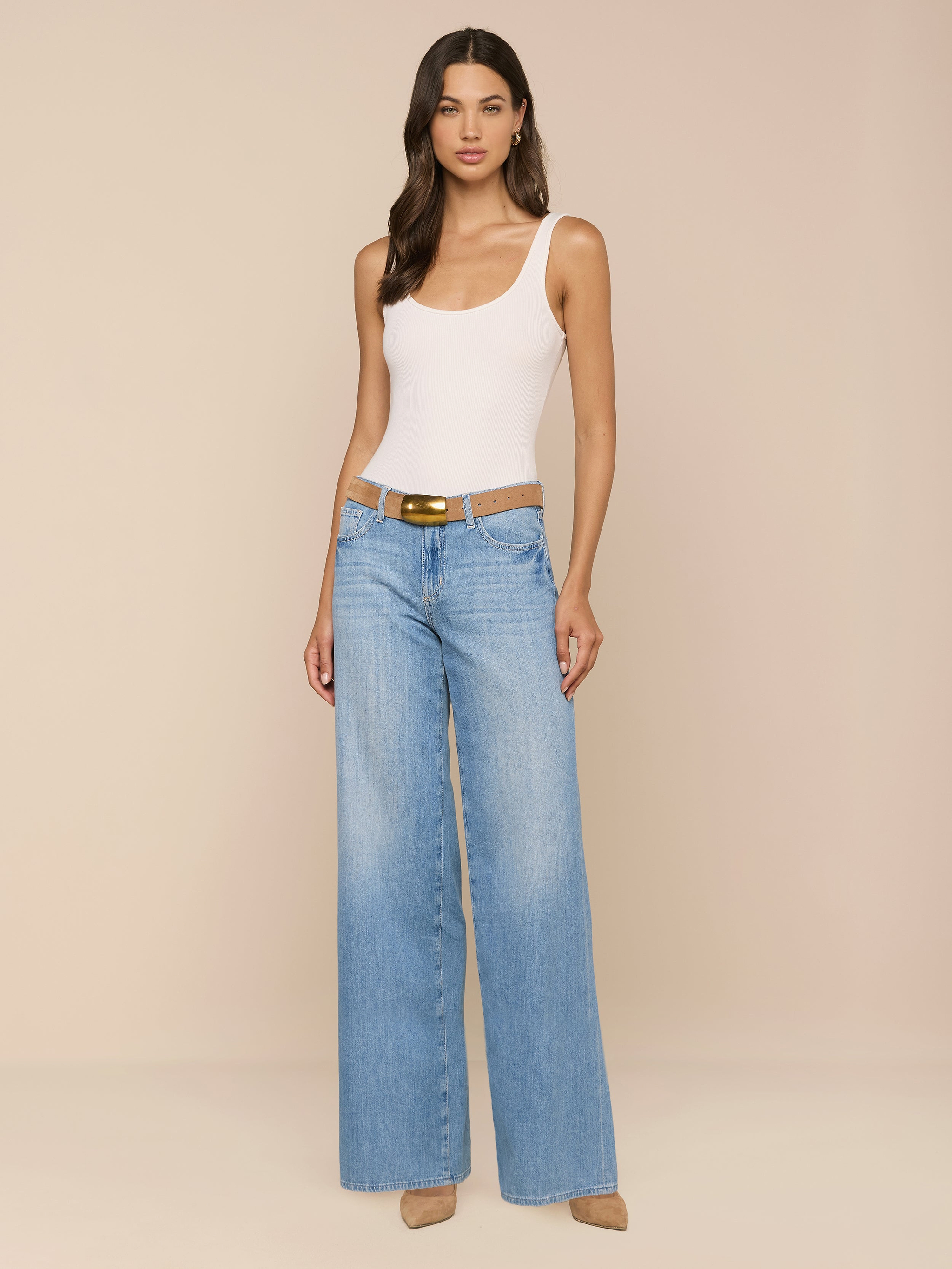 A woman stands against a plain white background, wearing a white tank top, the Alicent Wide-Leg Jean in light blue with a brown belt, and beige heels.