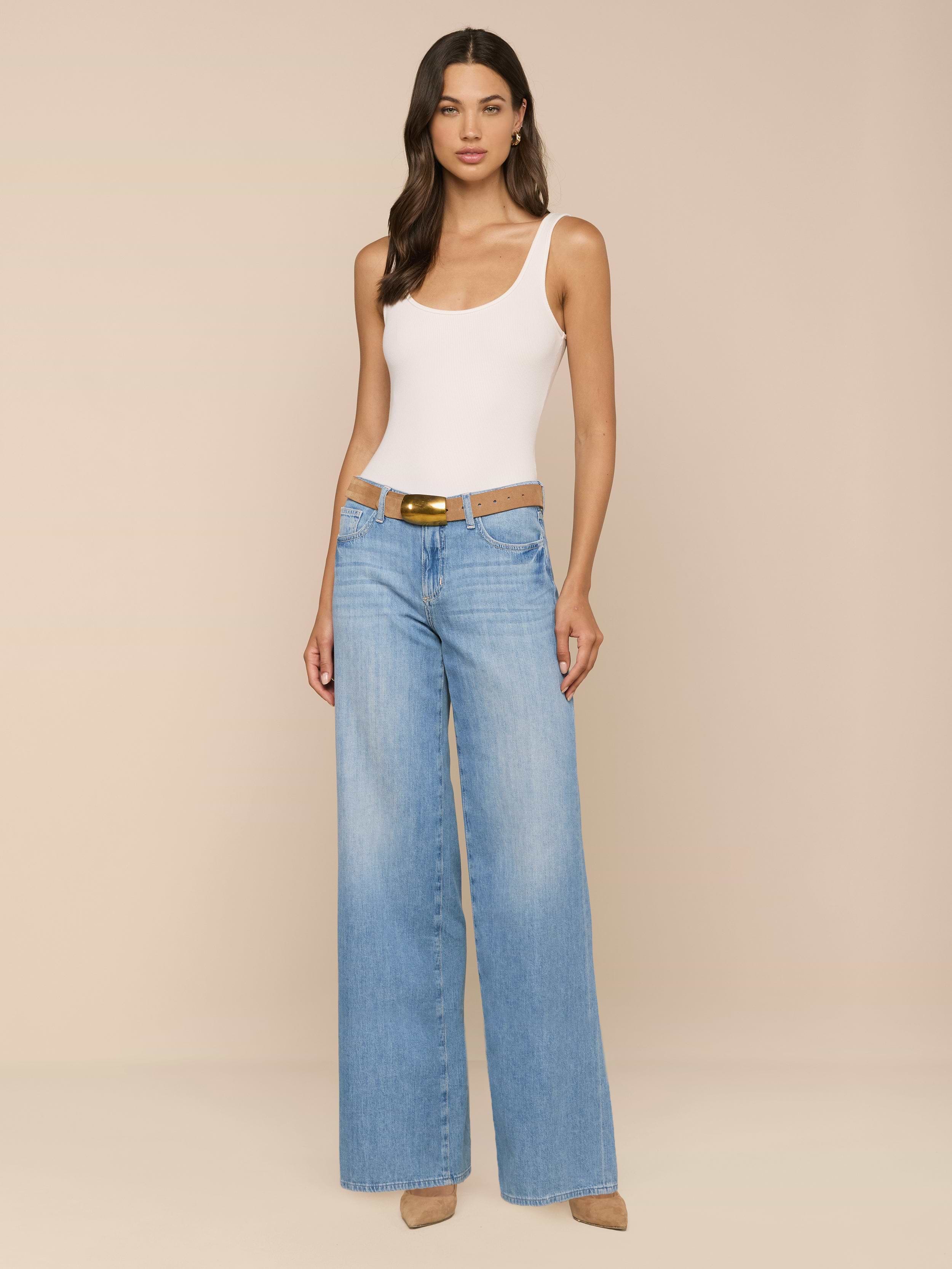 A woman stands against a plain white background, wearing a white tank top, the Alicent Wide-Leg Jean in light blue with a brown belt, and beige heels.