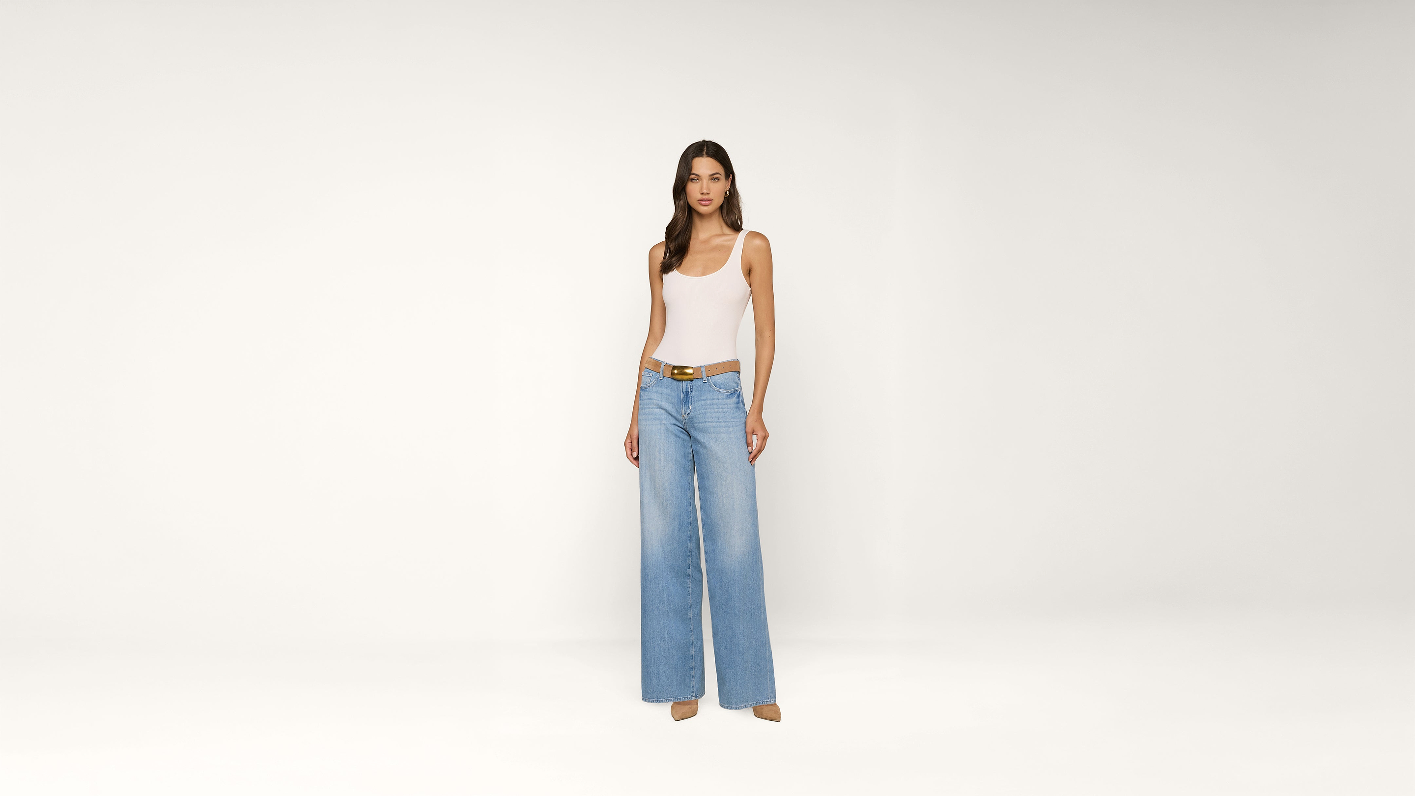 A woman stands against a plain white background, wearing a white tank top, light blue wide-leg jeans, a brown belt, and beige heels.