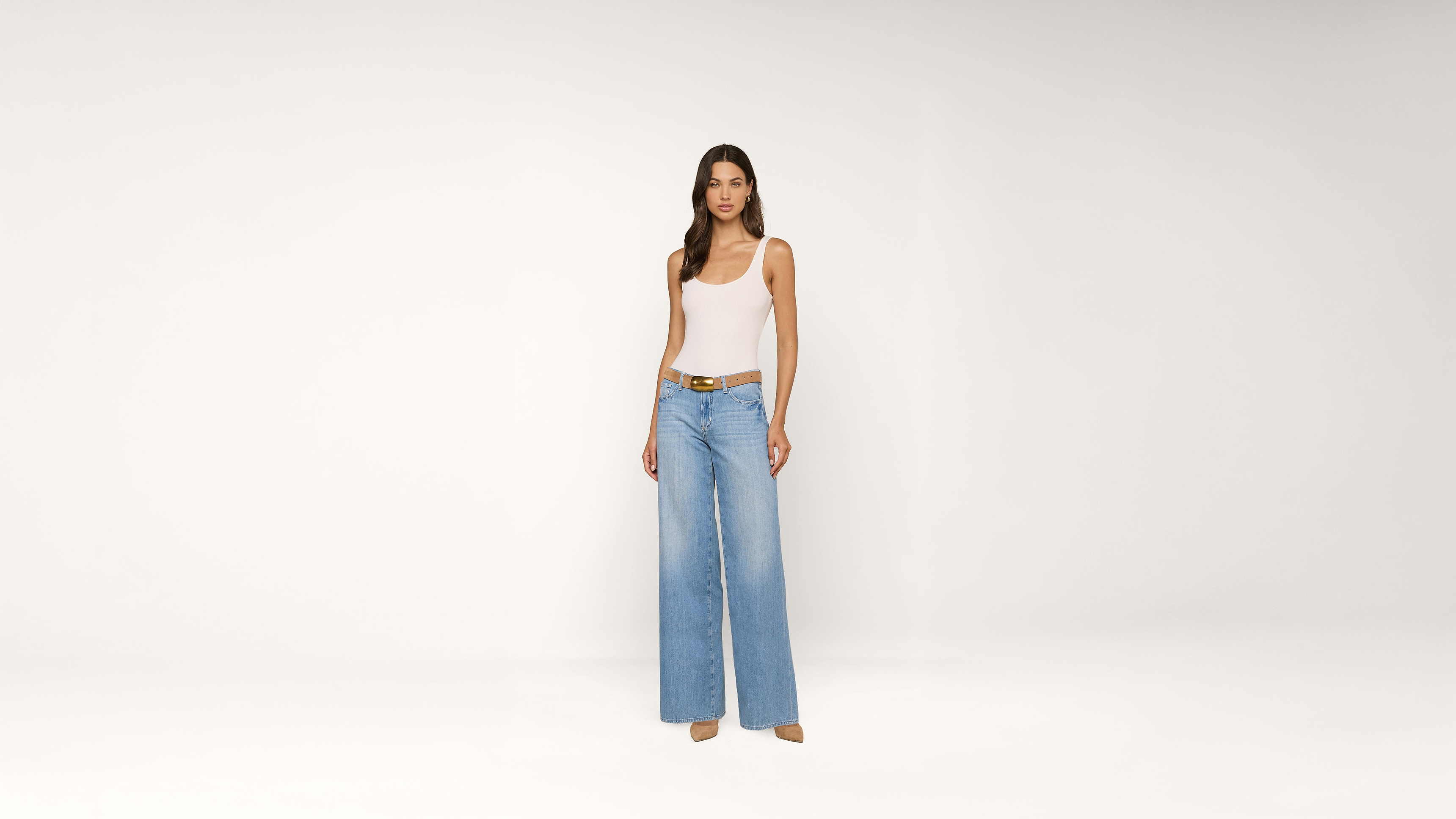A woman stands against a plain white background, wearing a white tank top, light blue wide-leg jeans, a brown belt, and beige heels.