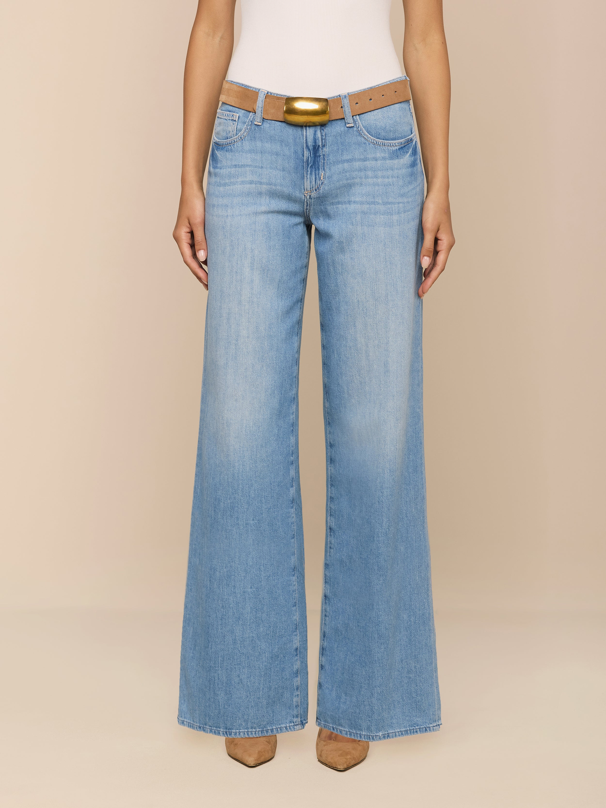 A person wearing the Alicent Wide-Leg Jean in light blue stands against a plain white background. The high rise, featherweight denim jeans feature a wide-leg silhouette and are styled with a brown belt and tan pointed-toe shoes. The upper body is cropped out of the frame.