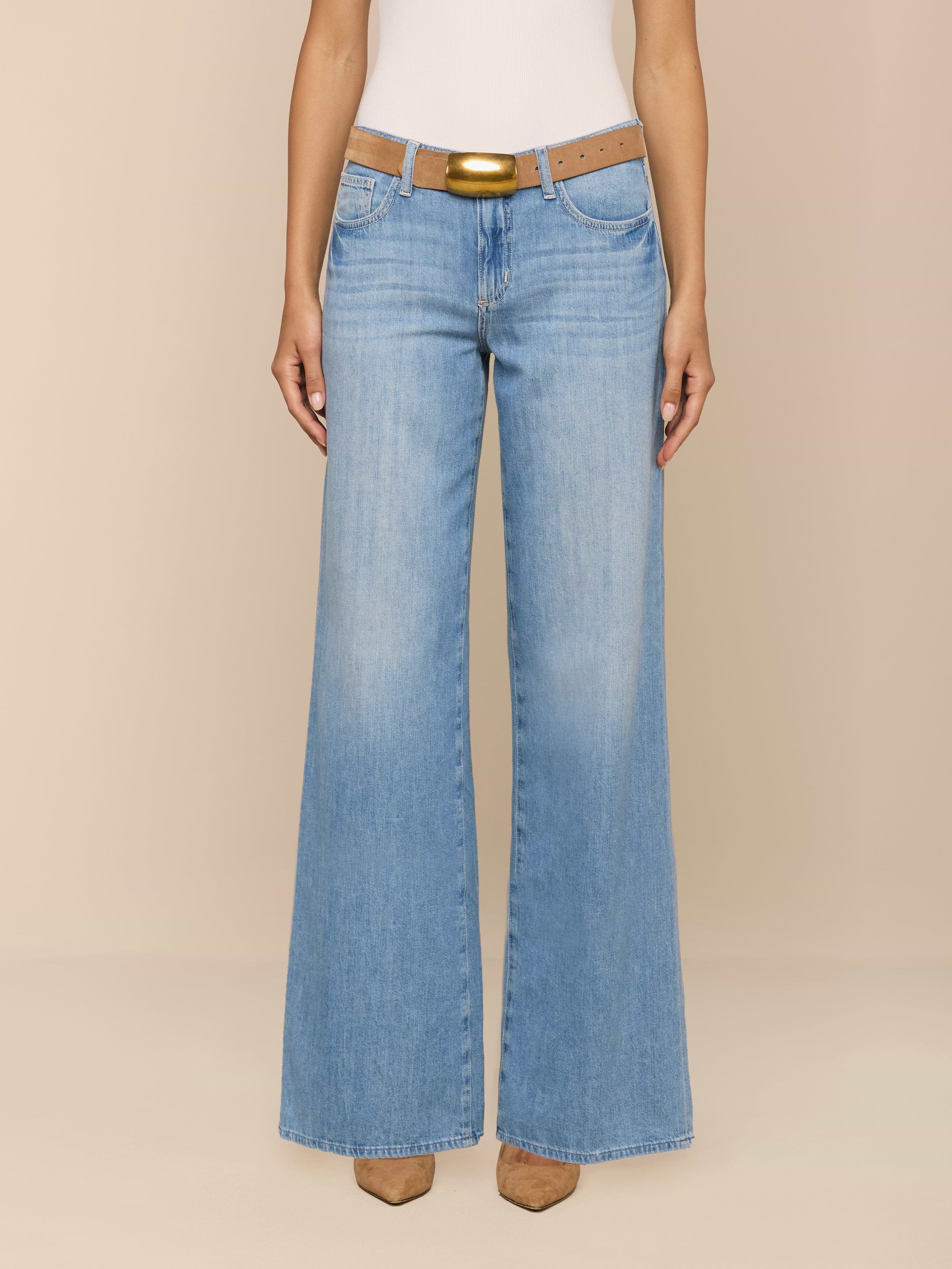 A person wearing the Alicent Wide-Leg Jean in light blue stands against a plain white background. The high rise, featherweight denim jeans feature a wide-leg silhouette and are styled with a brown belt and tan pointed-toe shoes. The upper body is cropped out of the frame.