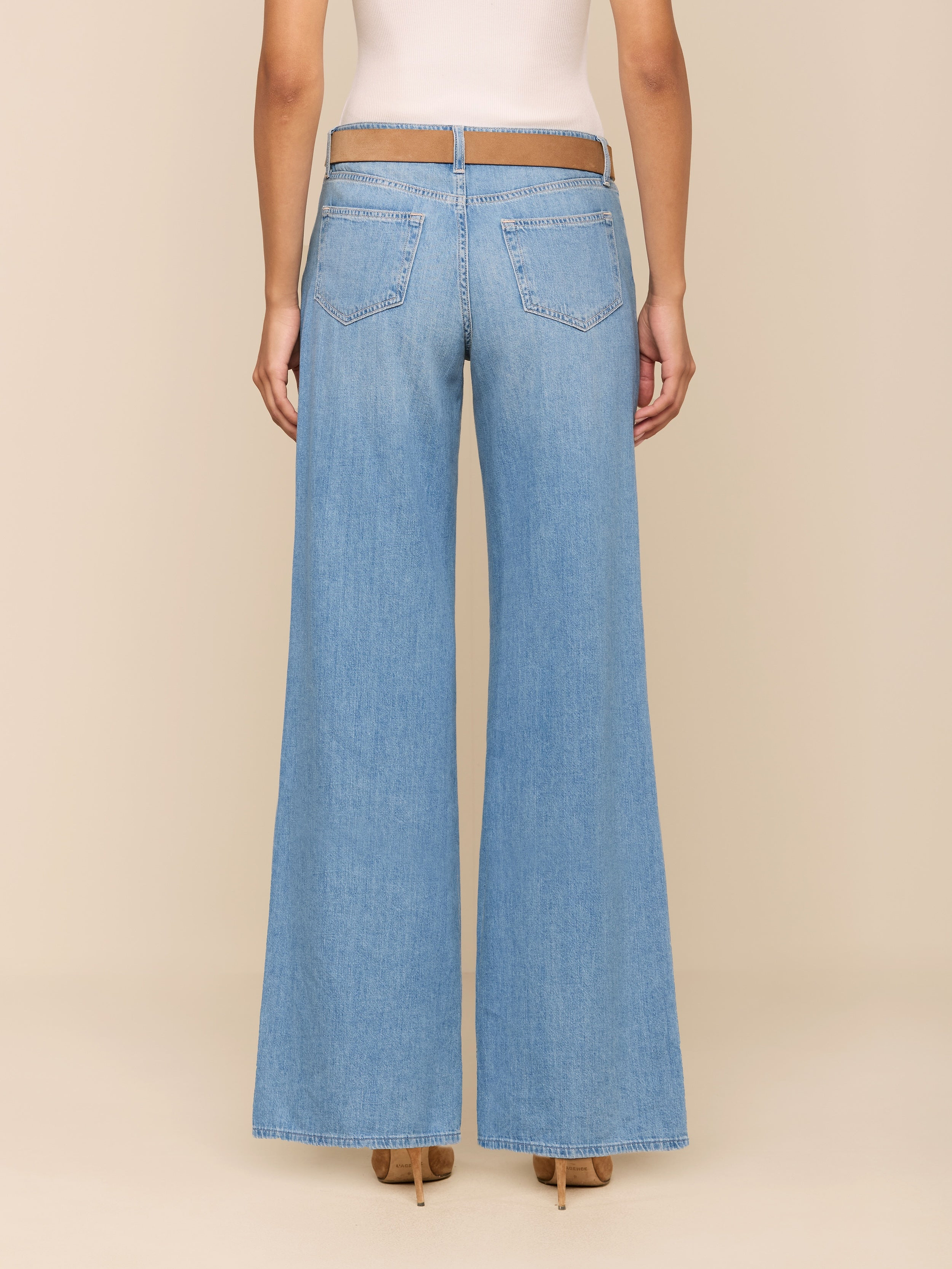 A person stands facing away against a plain white background, wearing the Alicent Wide-Leg Jean in light blue, styled with a tan belt, a light tank top, and tan heeled shoes.
