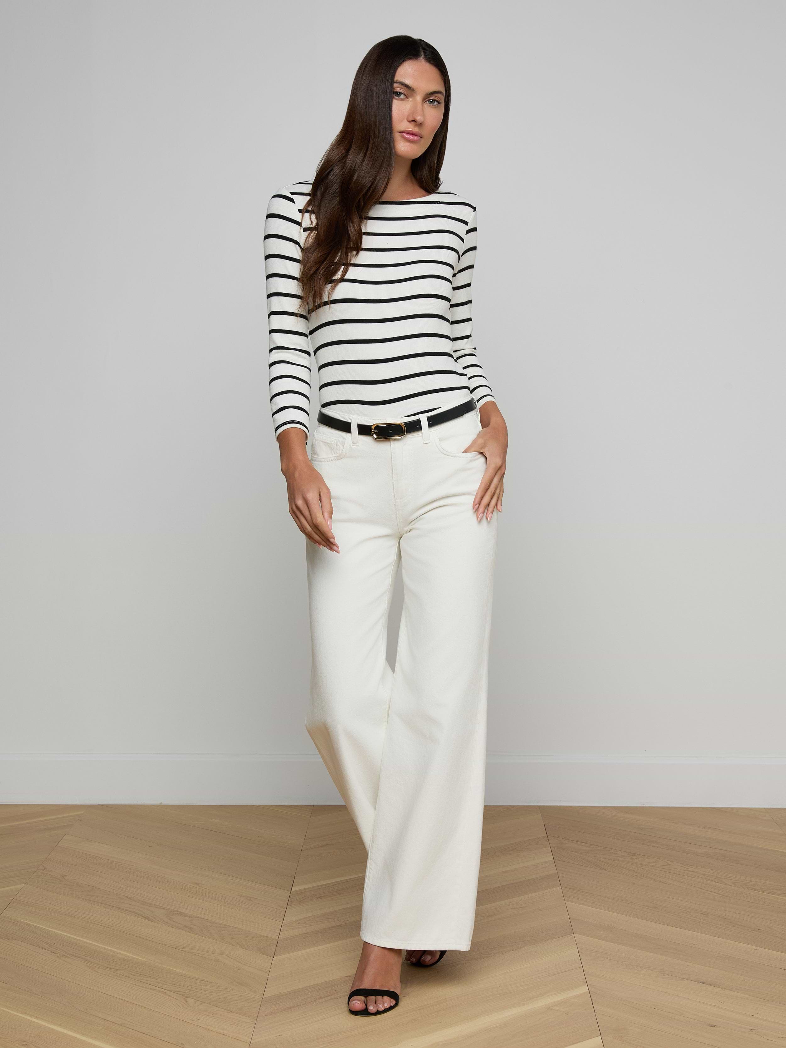 A woman stands against a plain background, wearing a black and white striped long-sleeve top, high rise Alicent Wide-Leg Jeans in white, a black belt, and black heels.
