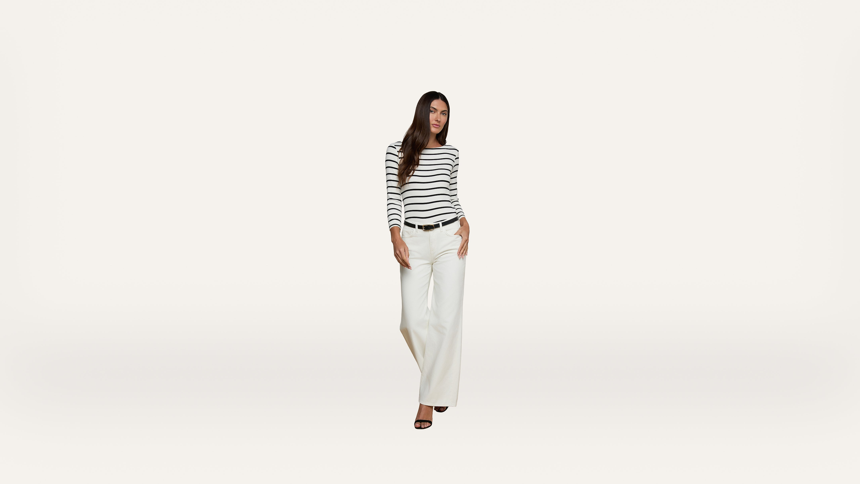 Woman wearing a black-and-white striped long-sleeve shirt, white wide-leg pants, and black heels stands against a plain white background.
