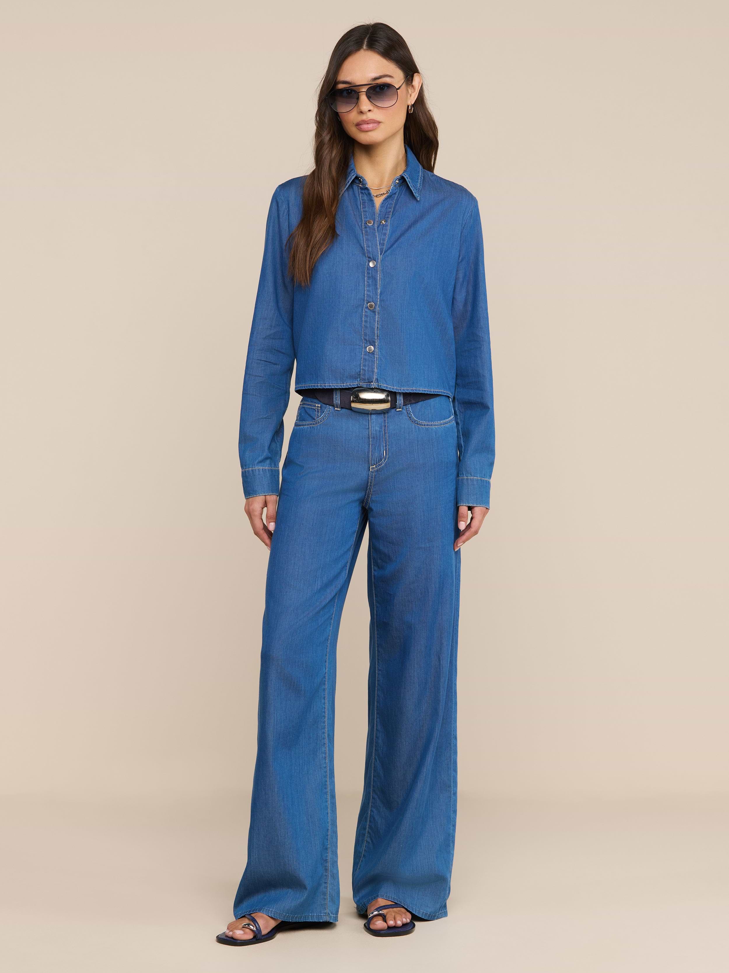 A woman wearing sunglasses, a blue featherweight denim button-up shirt, and the Alicent Wide-Leg Jean stands against a plain white background. She has long hair and is accessorized with sandals and a black belt.