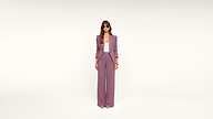 A person stands against a plain white background, wearing a mauve suit with wide-legged pants, a matching blazer, a light top, sunglasses, and beige heels.