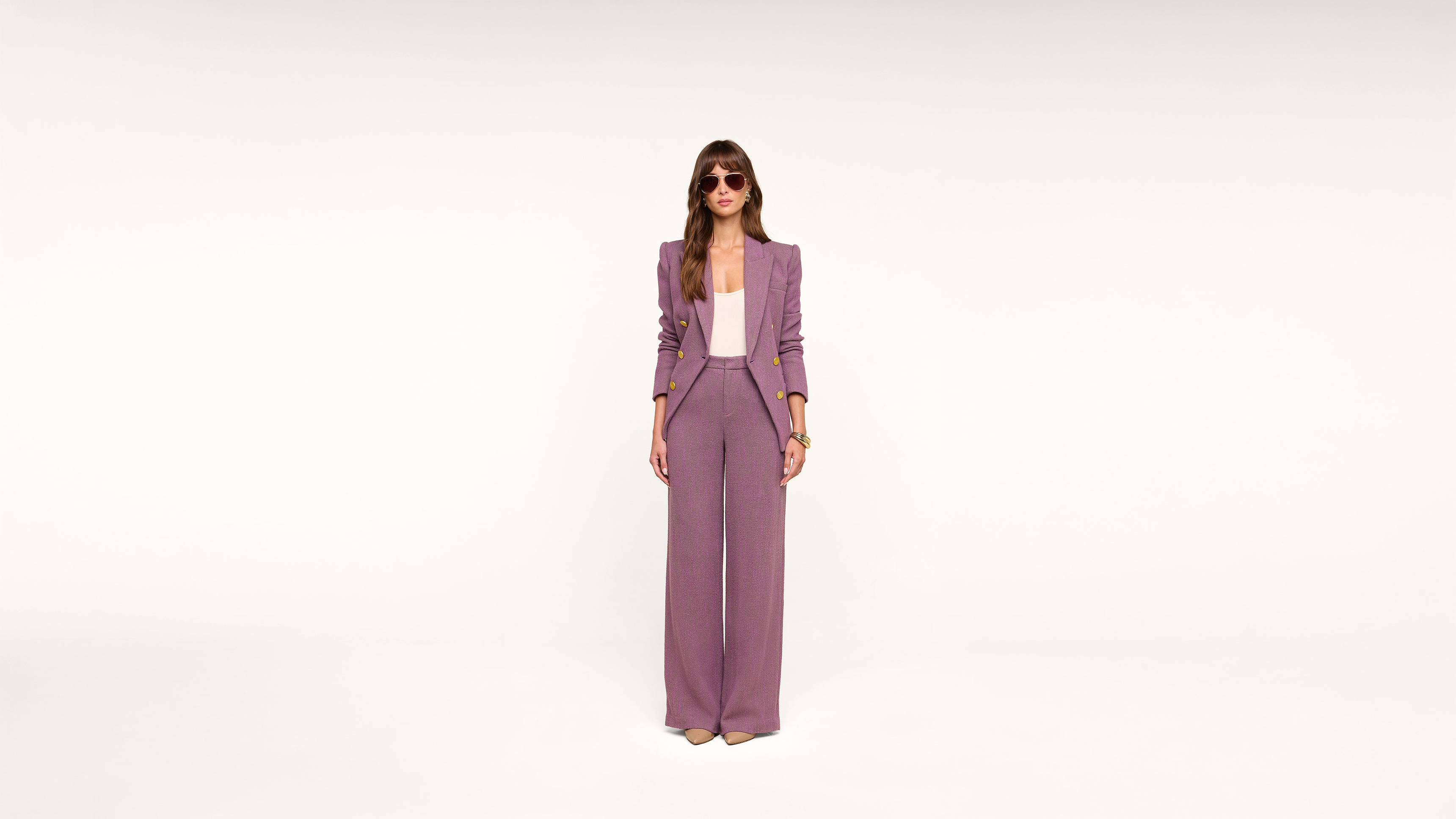 A person stands against a plain white background, wearing a mauve suit with wide-legged pants, a matching blazer, a light top, sunglasses, and beige heels.