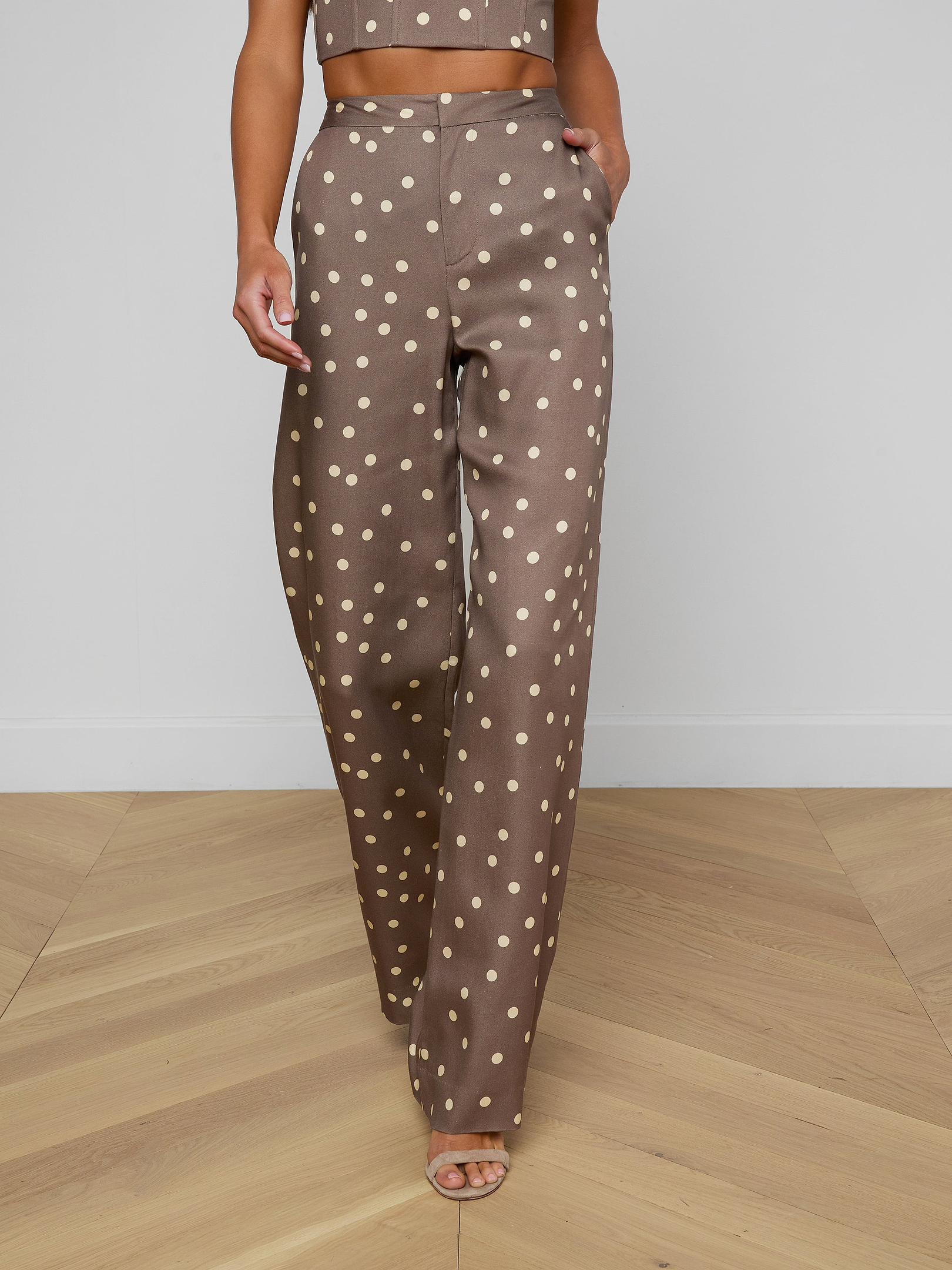 Wearing the Livvy Straight-Leg Trouser featuring a brown polka dot print, a model walks forward against a plain background, styled with a matching crop top and open-toe heels.
