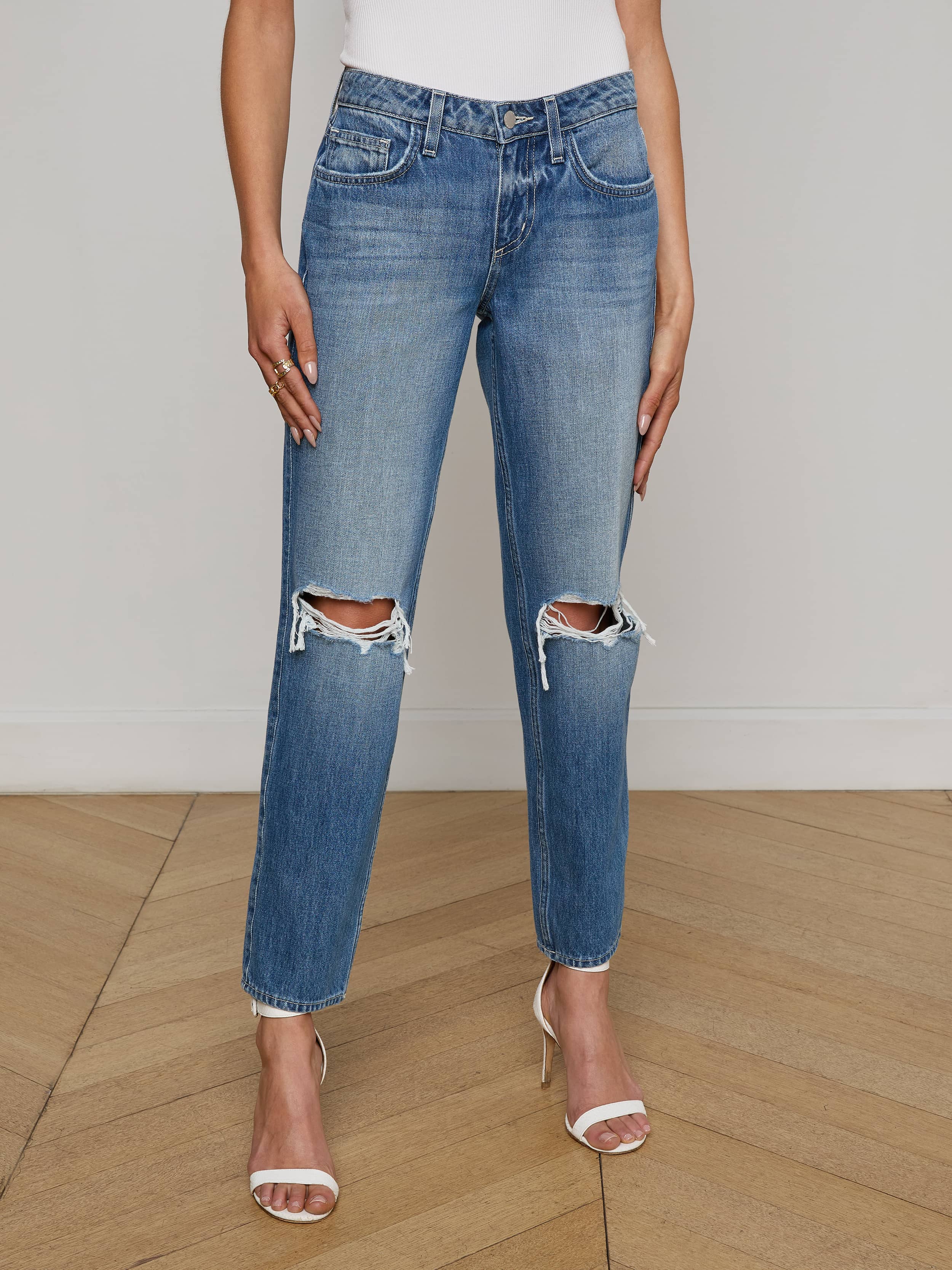 Wearing the Nevia Slouchy Straight-Leg Jean in a blue wash with ripped knees, paired with a white top and nude high-heeled sandals, this person stands against a plain white background.