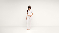A woman wearing sunglasses, a white cropped shirt, white pants, and white sandals stands against a plain white background with a relaxed posture.