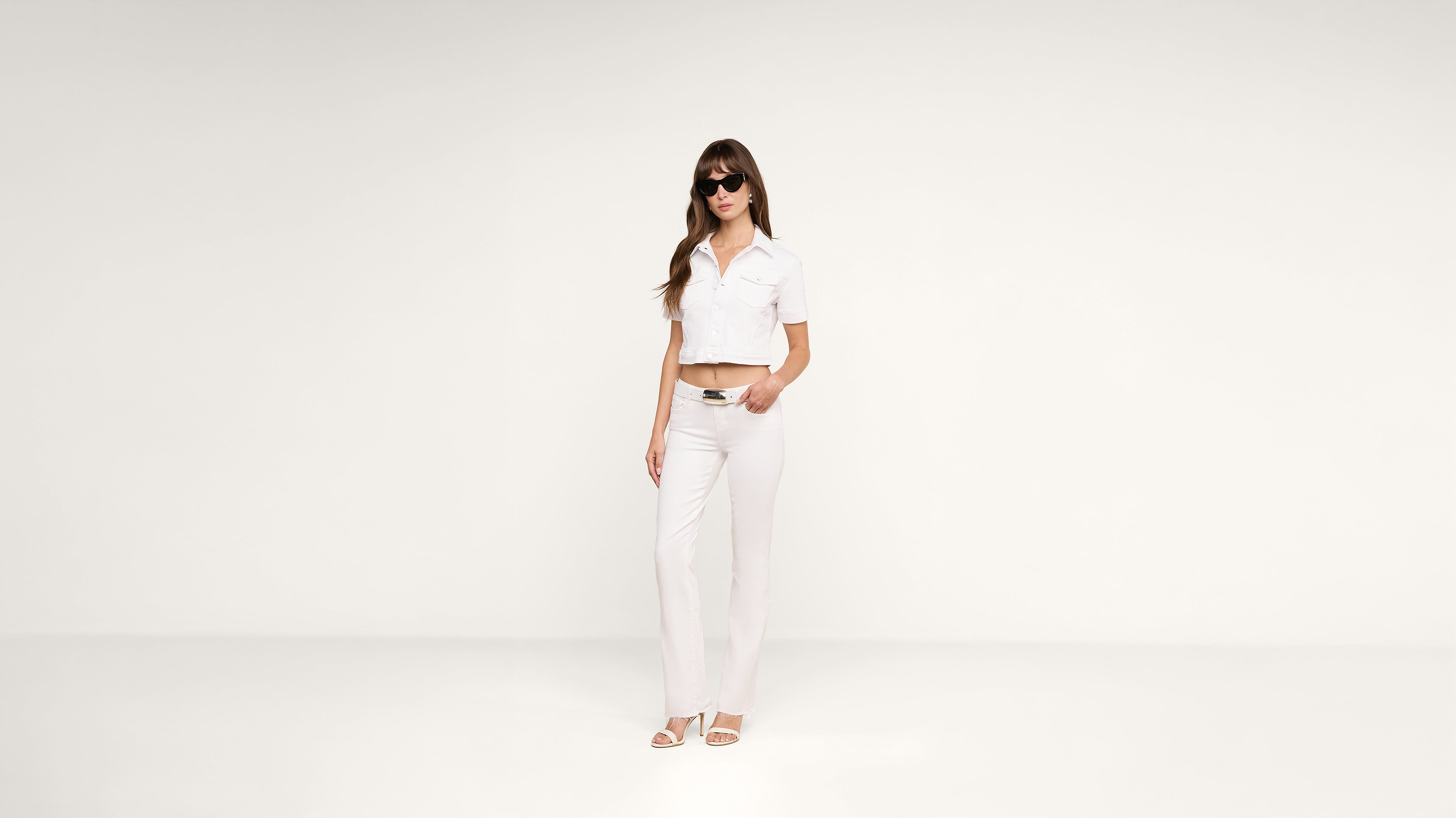 A woman wearing sunglasses, a white cropped shirt, white pants, and white sandals stands against a plain white background with a relaxed posture.