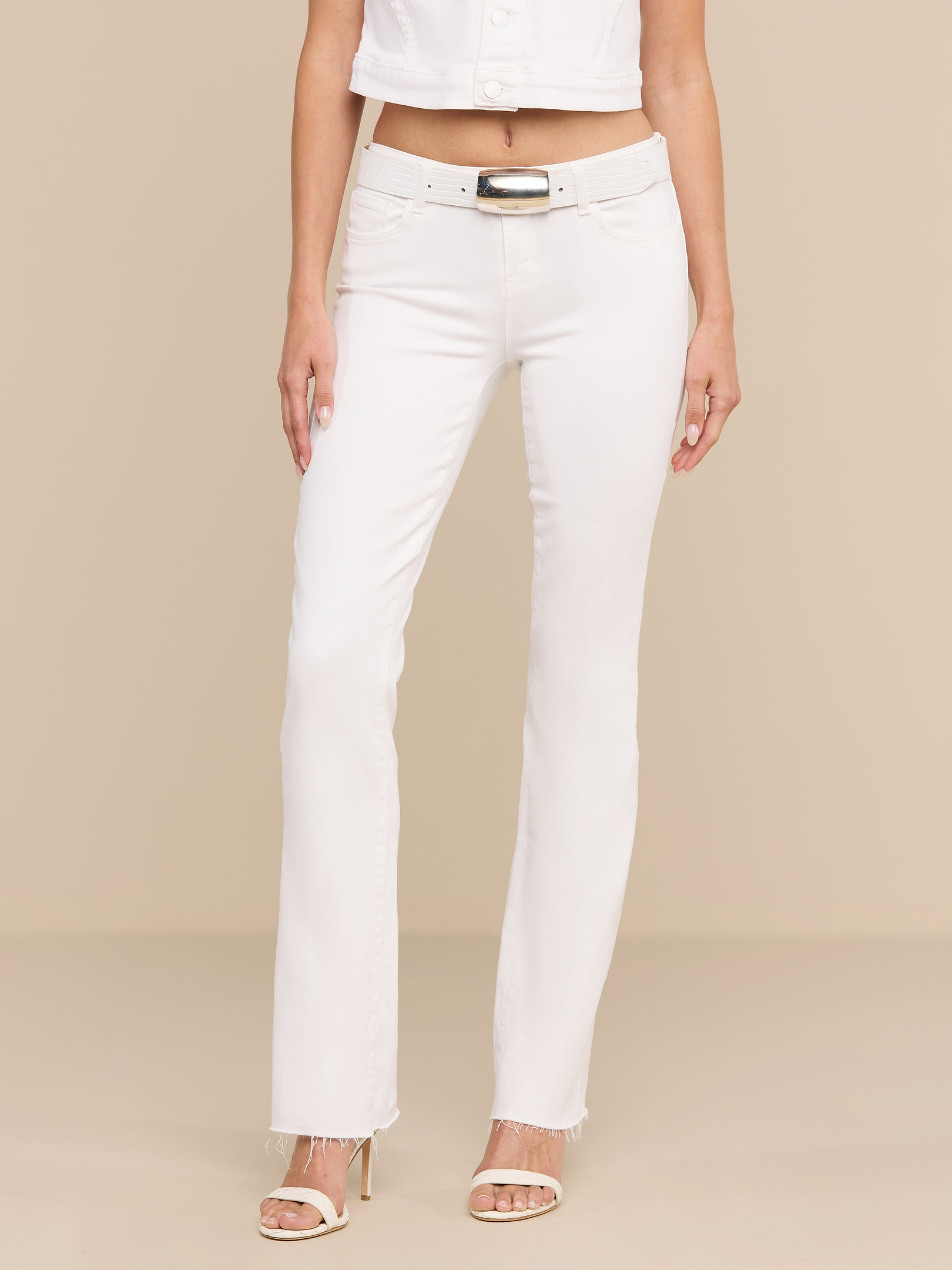 Une personne porte un jean « Sneeki » taille basse à coupe droite, avec une ceinture blanche, des sandales blanches à talons hauts et une veste blanche courte, debout devant un fond beige.