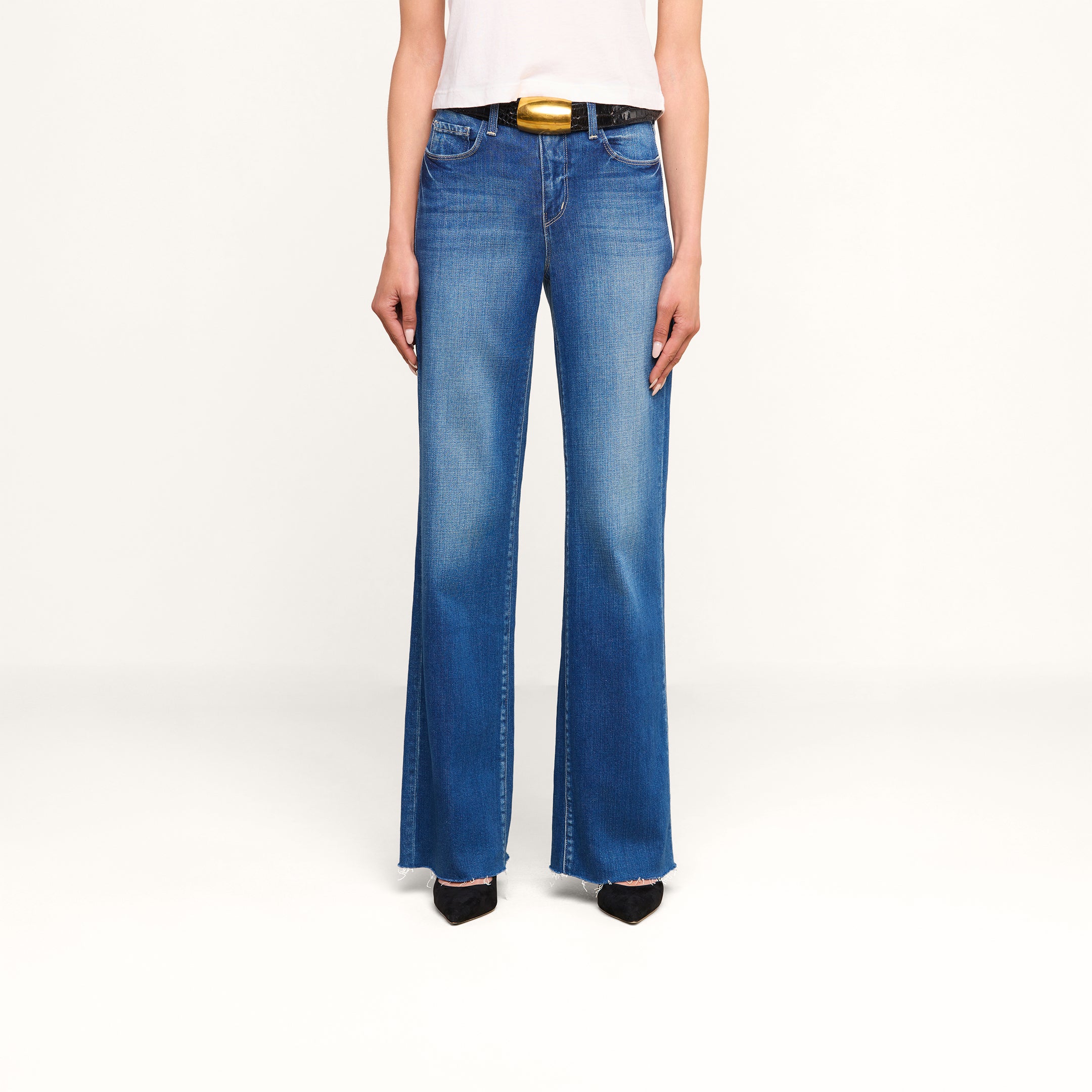 Una modella indossa i jeans Scottie Wide-Leg in denim blu con lavaggio vintage e orli sfrangiati, abbinati a una cintura nera con una grande fibbia dorata, un top bianco e scarpe nere a punta, in piedi su uno sfondo beige a tinta unita.