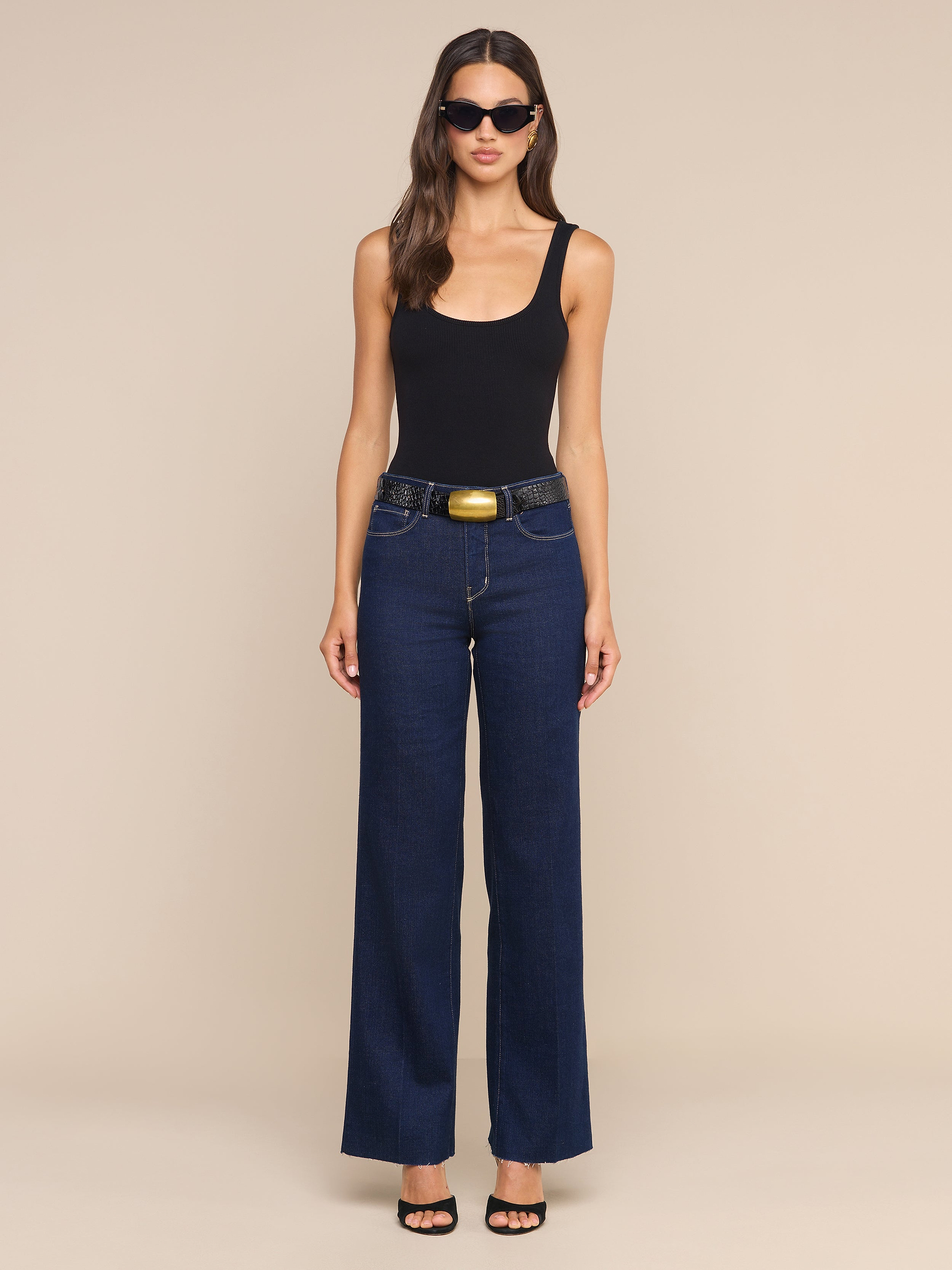 Una donna è in piedi su uno sfondo beige, con indosso una canotta nera, jeans Scottie Wide-Leg realizzati in denim elasticizzato di alta qualità, una cintura nera, sandali neri, occhiali da sole e capelli sciolti e mossi.