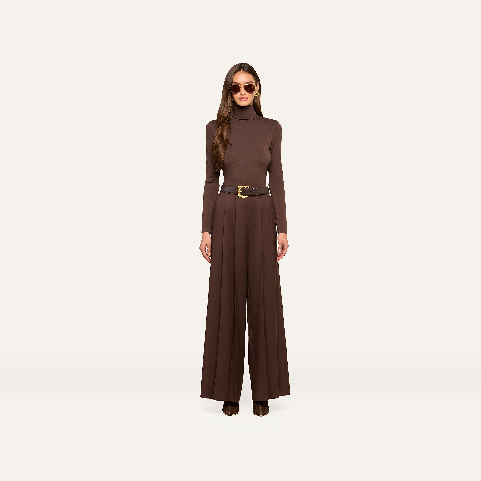 A woman wearing sunglasses, a brown long-sleeve turtleneck top, the Nova Pleated Wide-Leg Pant with a belt in fluid suiting fabric, and brown shoes stands facing forward against a plain white background.