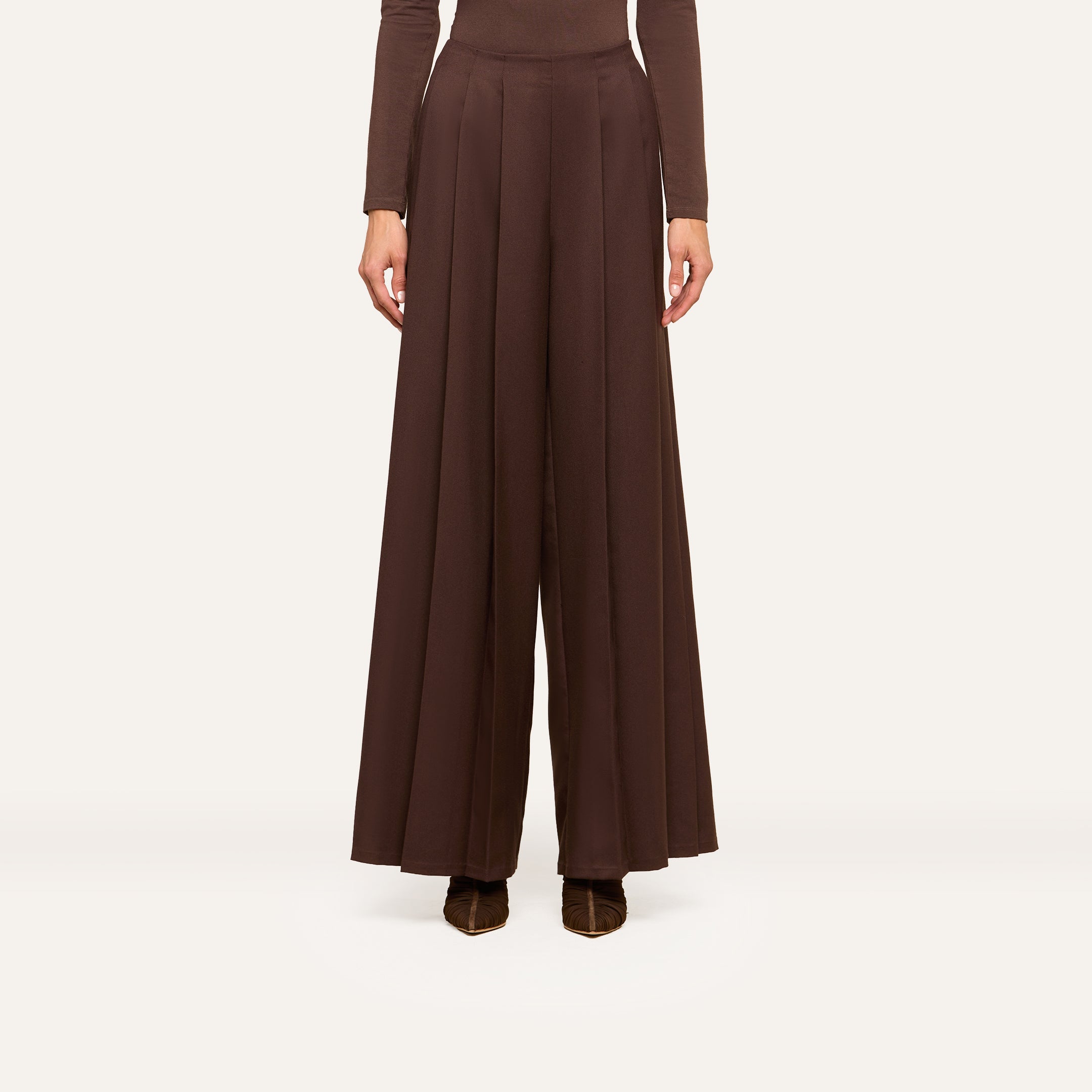 A person wearing ultra high rise Nova Pleated Wide-Leg Pants in brown, crafted from fluid suiting fabric, and a coordinating brown long-sleeve top stands against a plain light background.