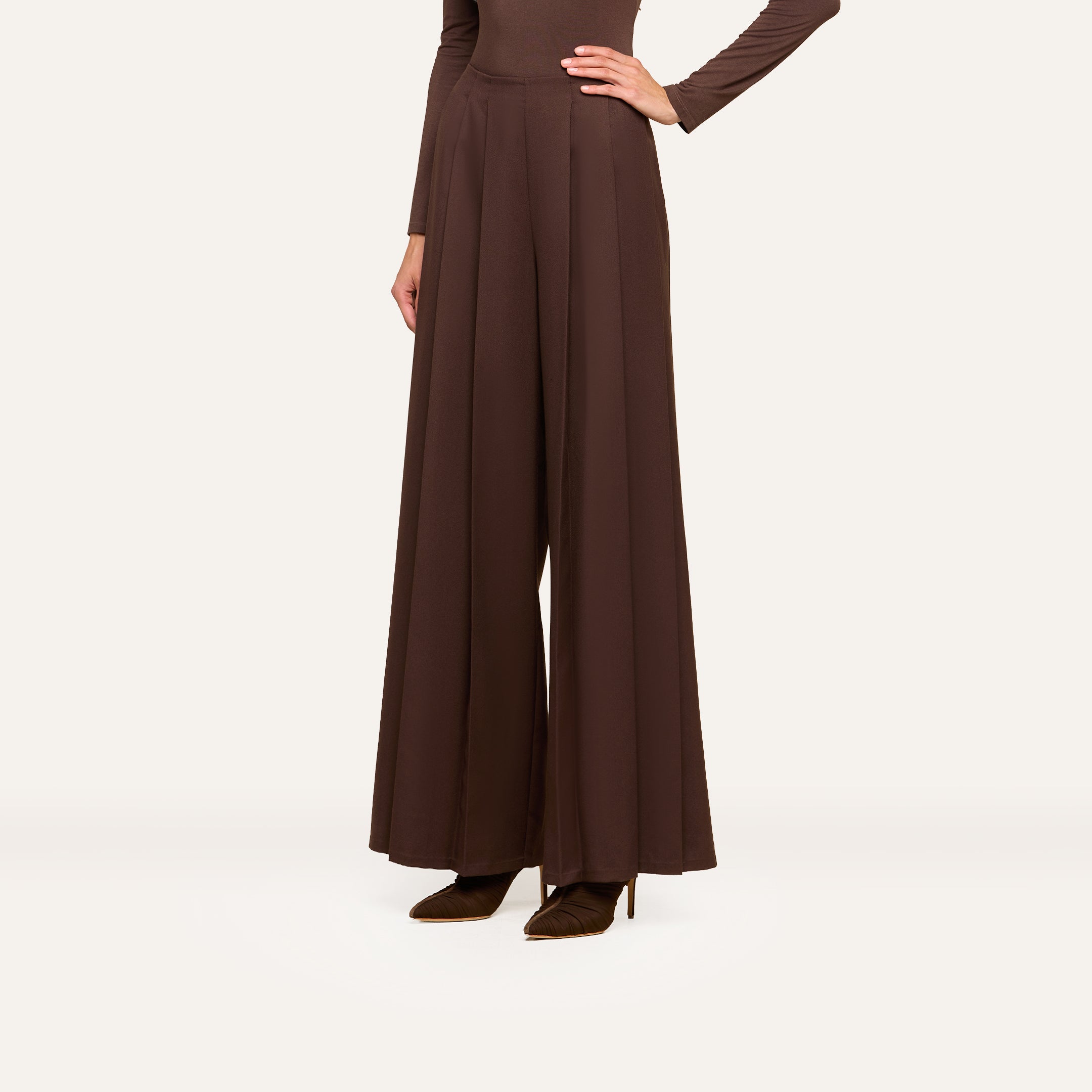 A person wearing ultra high rise, wide-leg Nova Pleated Wide-Leg Pants in brown fluid suiting fabric and pointed brown heels, with one hand on their hip, stands against a plain light background.