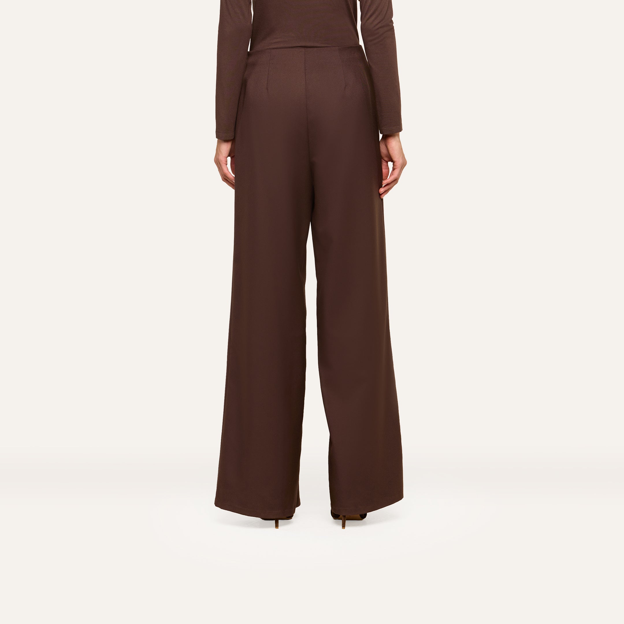 Wearing the Nova Pleated Wide-Leg Pant, crafted from a fluid suiting fabric in an ultra high rise and wide-leg silhouette, a person in a matching long-sleeve top stands facing away against a plain, light background.