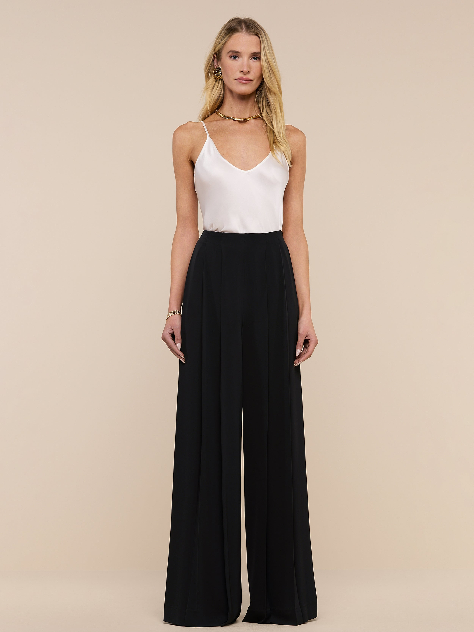 A woman stands against a plain white background, wearing a white sleeveless top paired with the Nova Pleated Wide-Leg Pant, a high-waisted tailored black trouser. Her outfit features wide-leg trousers. She has long blonde hair and a neutral expression.