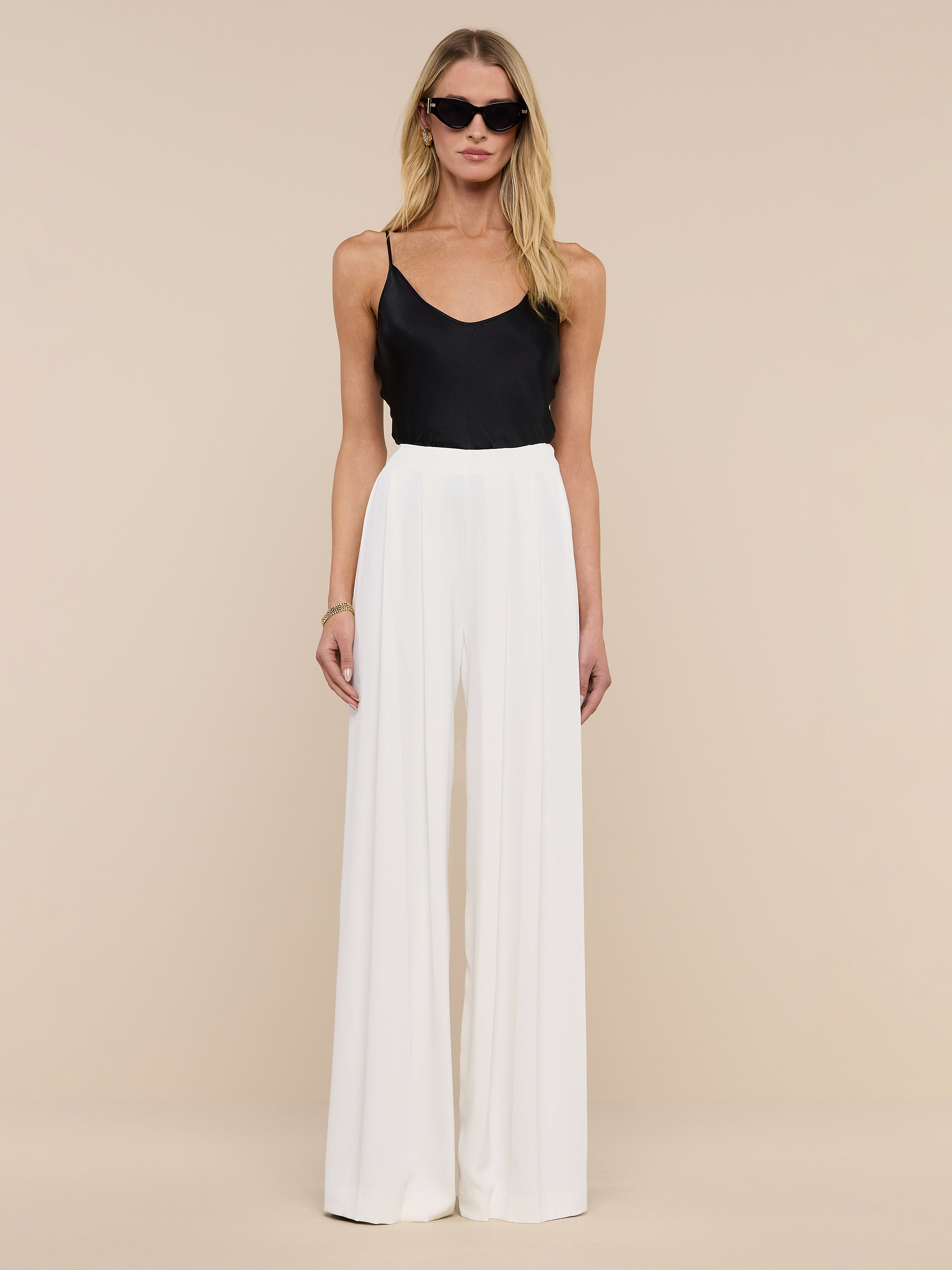 A woman in sunglasses wears a black sleeveless top and the Nova Pleated Wide-Leg Pant in ivory, standing against a plain white background.