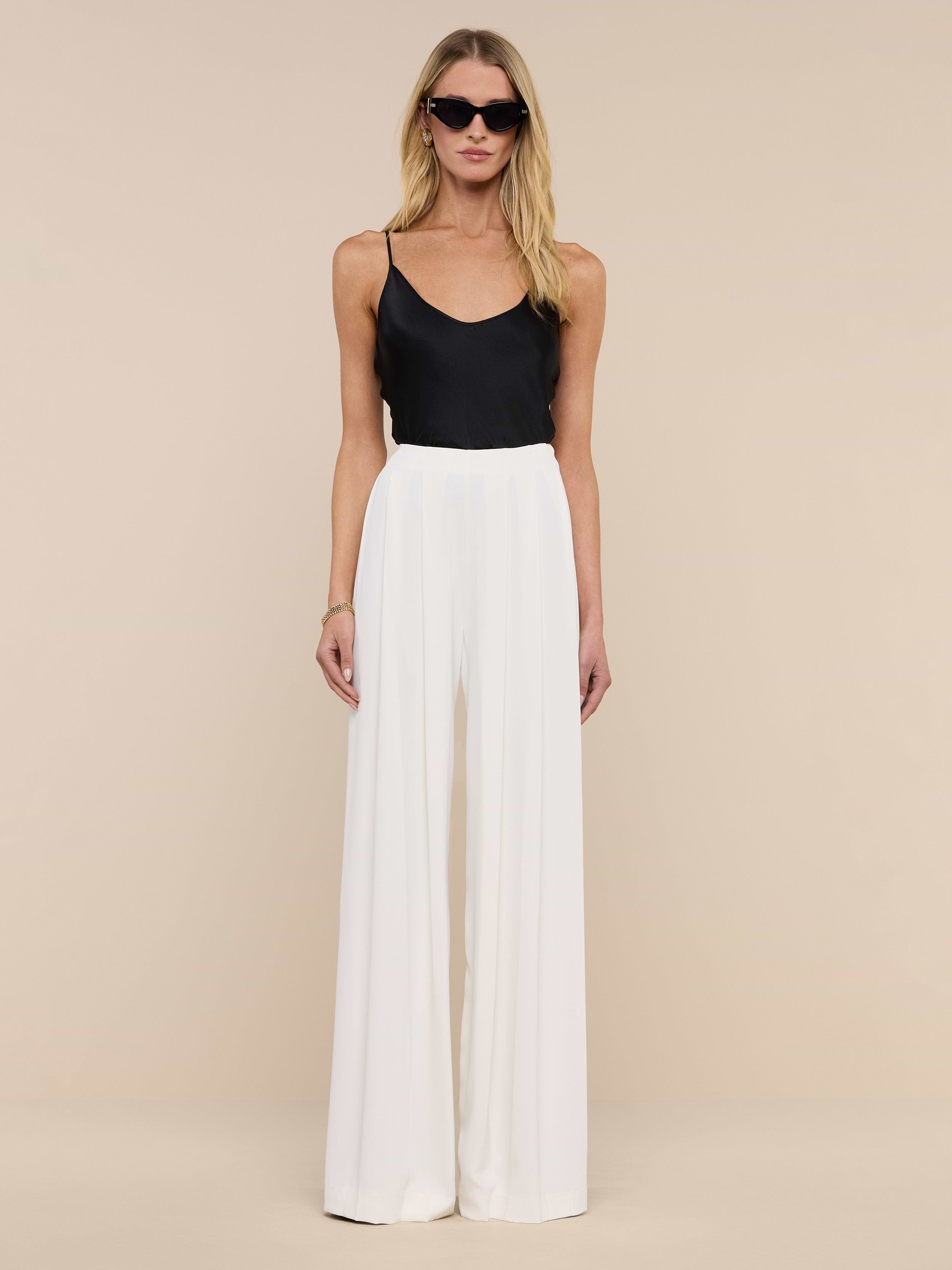A woman in sunglasses wears a black sleeveless top and the Nova Pleated Wide-Leg Pant in ivory, standing against a plain white background.