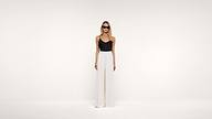 Woman wearing sunglasses, a black sleeveless top, and high-waisted wide-leg white pants stands against a plain white background, looking straight ahead with her arms relaxed by her sides.