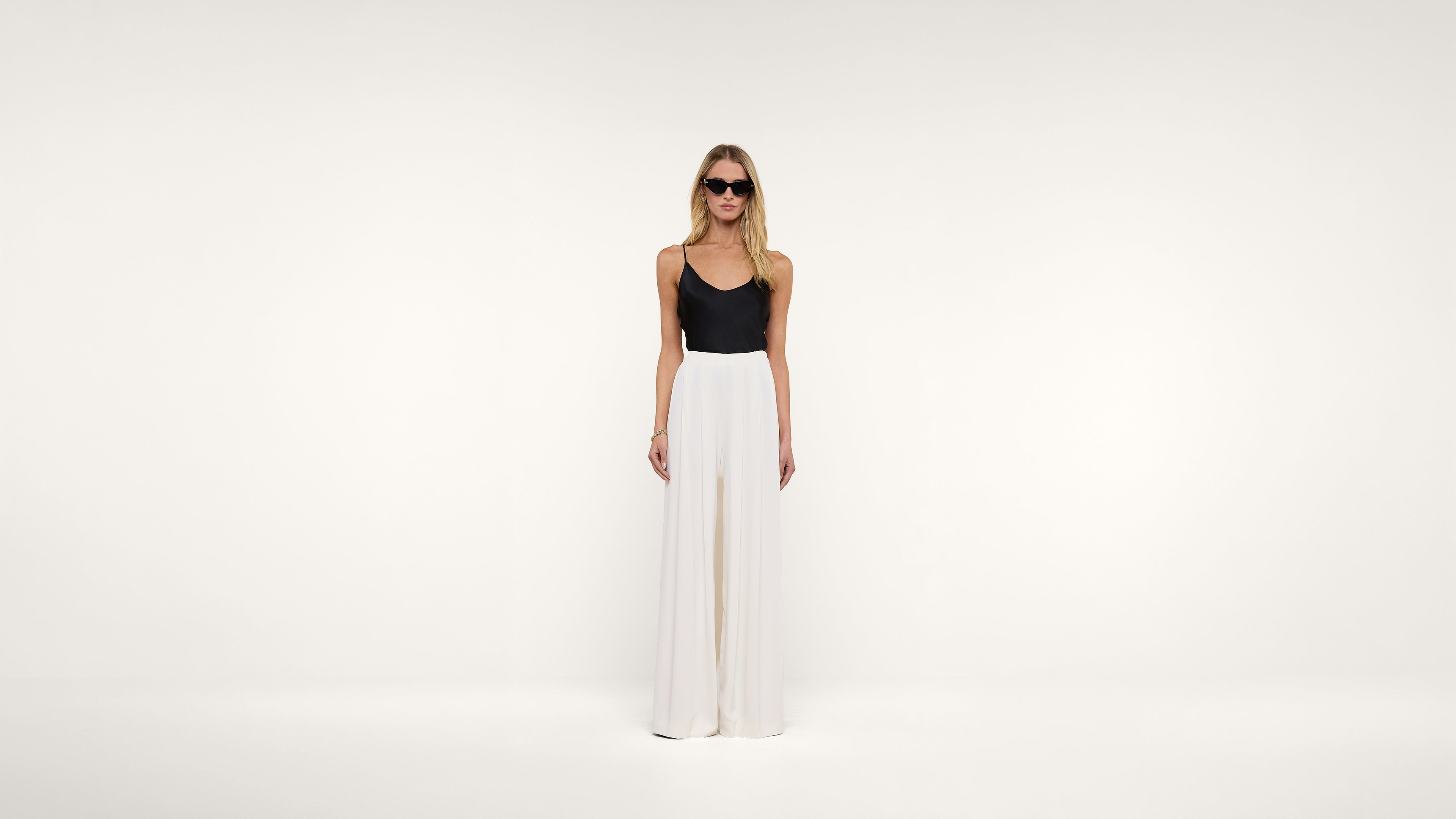 Woman wearing sunglasses, a black sleeveless top, and high-waisted wide-leg white pants stands against a plain white background, looking straight ahead with her arms relaxed by her sides.